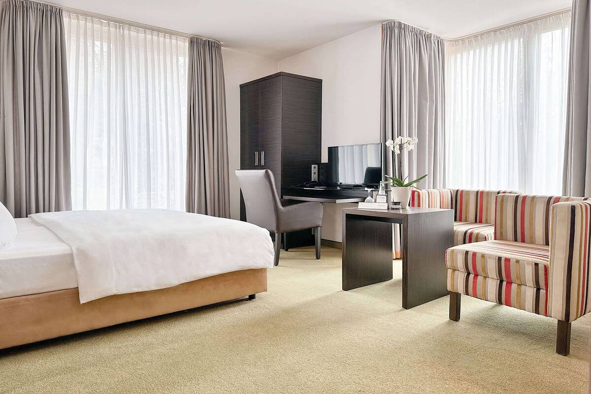 Una habitación de hotel con cama, escritorio, sillón a rayas y un armario de color oscuro.