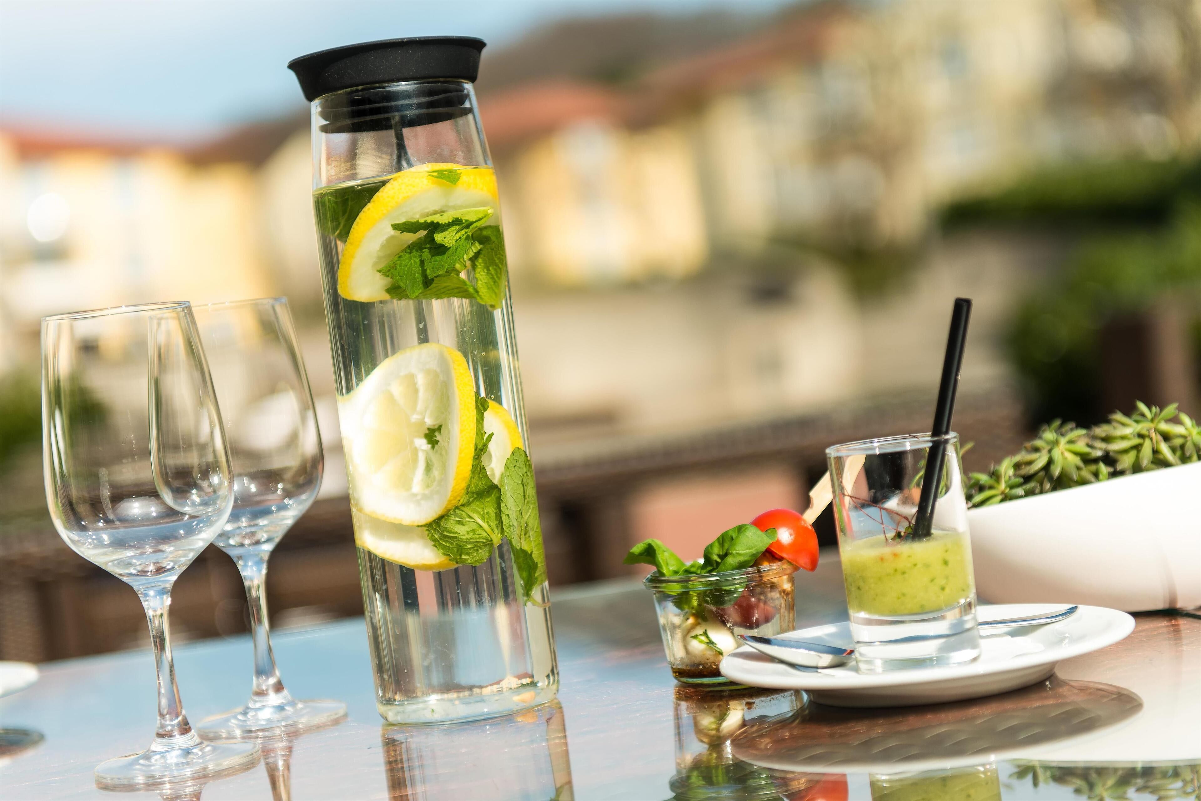 A table with a tall glass of water with lemon slices and mint, two empty wine glasses, and a small dish with a green drink and a cherry.