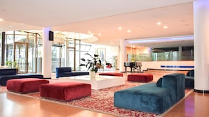 Un hall d'entrée moderne avec des sièges rouges et bleus, une table blanche au centre agrémentée d'une plante, et un piano en arrière-plan.