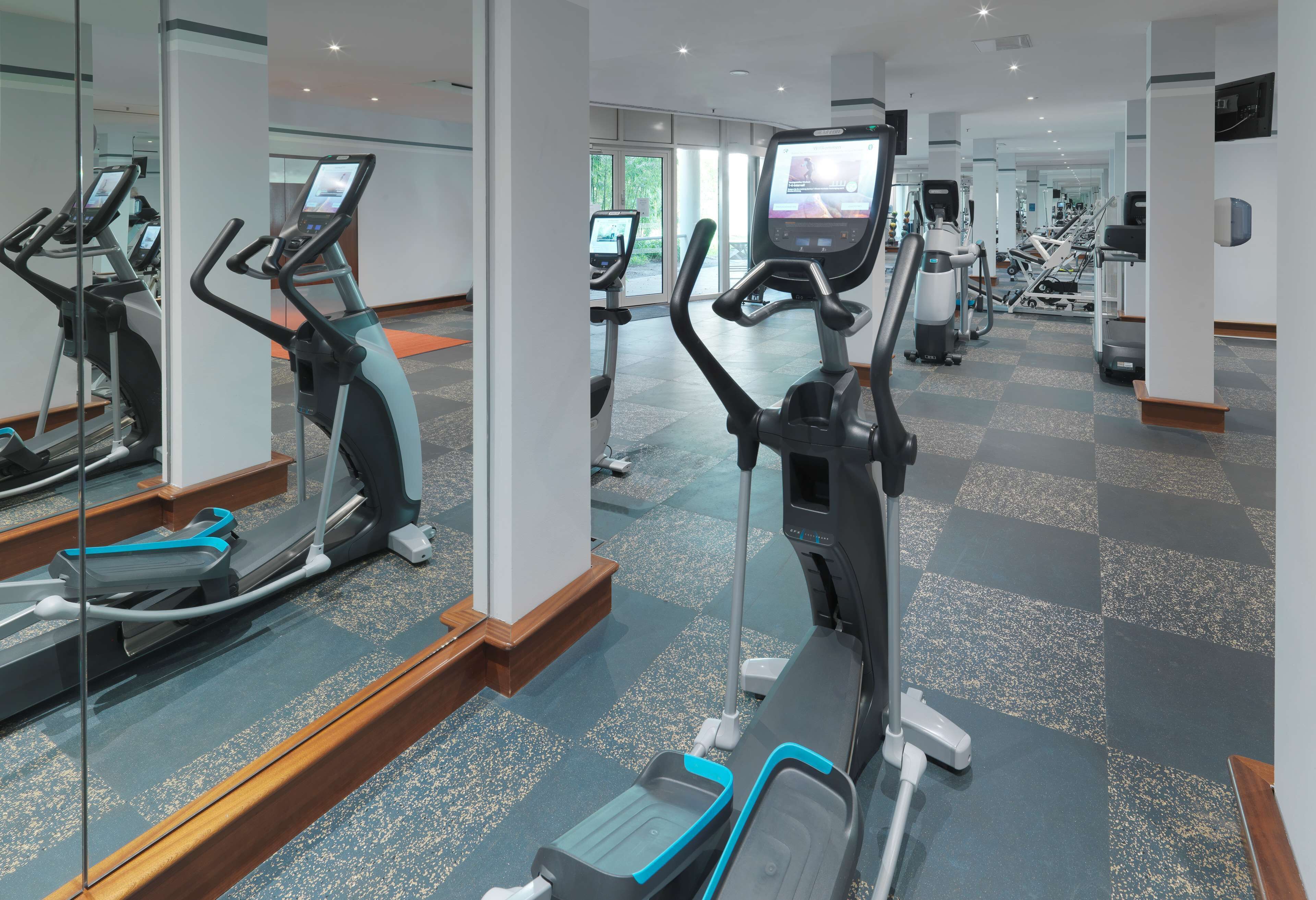 A modern gym with multiple cardio machines, mirrored walls, and a checkered floor.