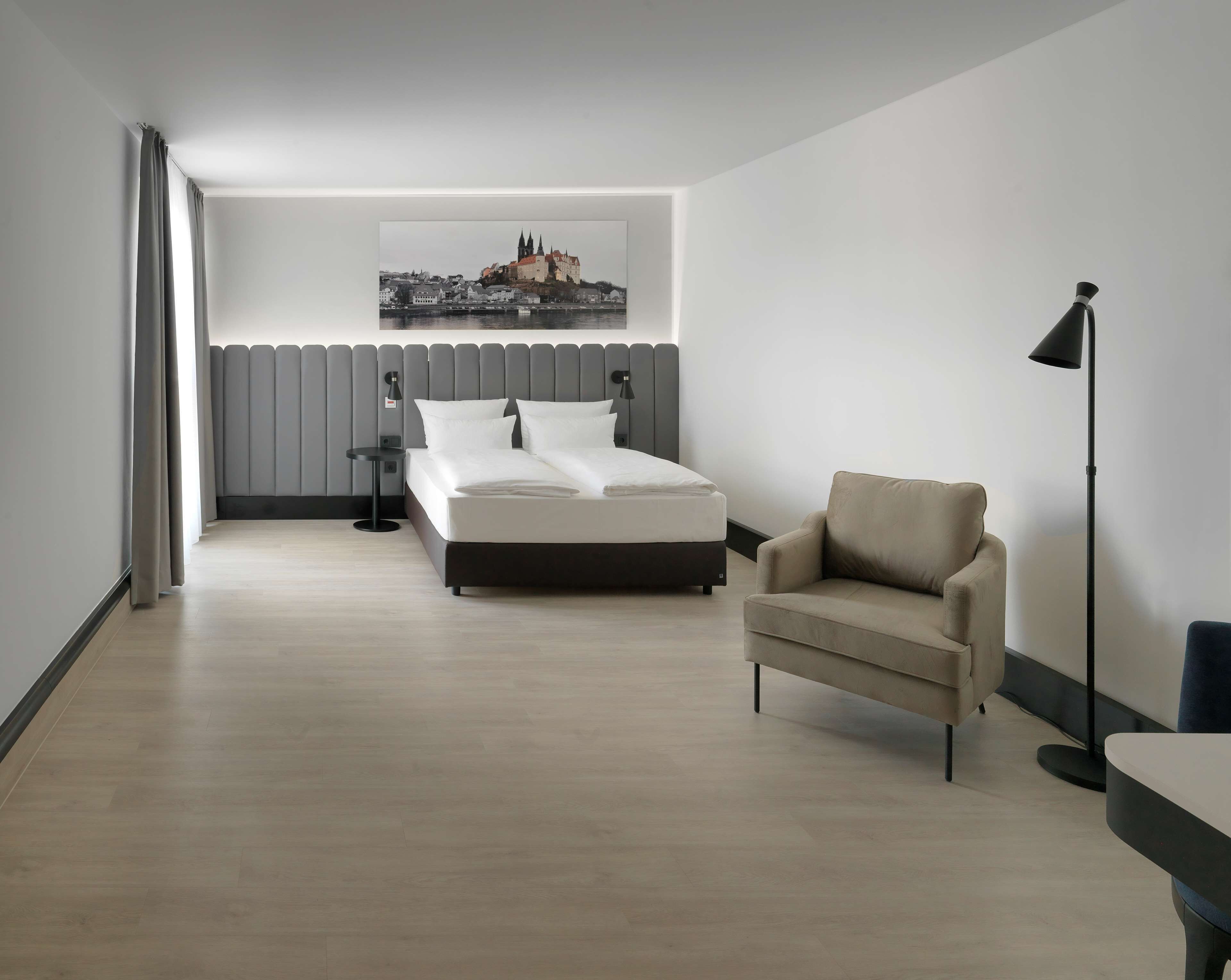 Et moderne hotelværelse med seng, stol, skrivebord, lampe og et bybillede på væggen.