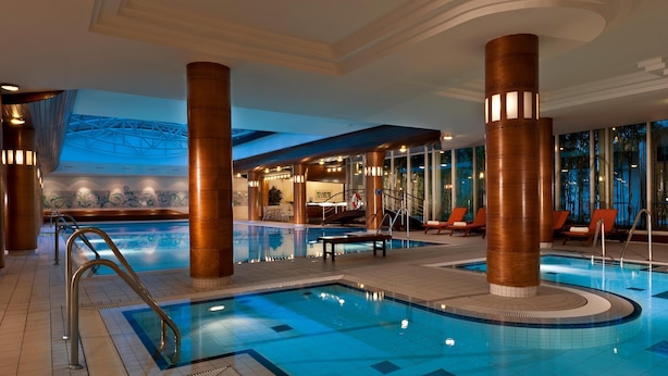 Une grande piscine intérieure avec un sol carrelé, entourée de chaises longues et d’un escalier menant à un niveau supérieur.