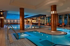 Una gran piscina interior con suelo de baldosas, rodeada de sillones de descanso y una escalera que lleva a un nivel superior.