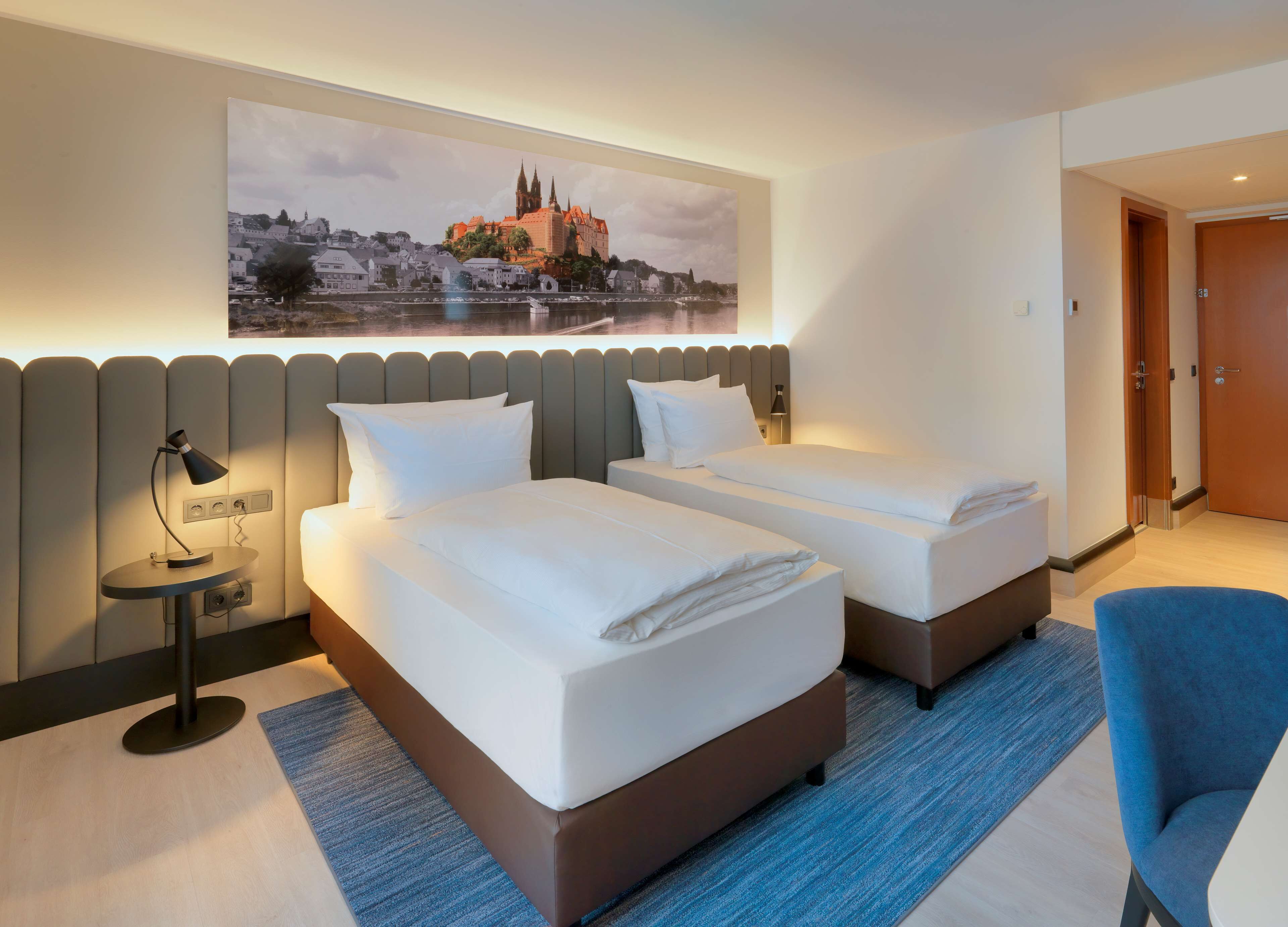 A hotel room with two beds, a nightstand with a lamp, a large window with a cityscape view, and a door to the right.