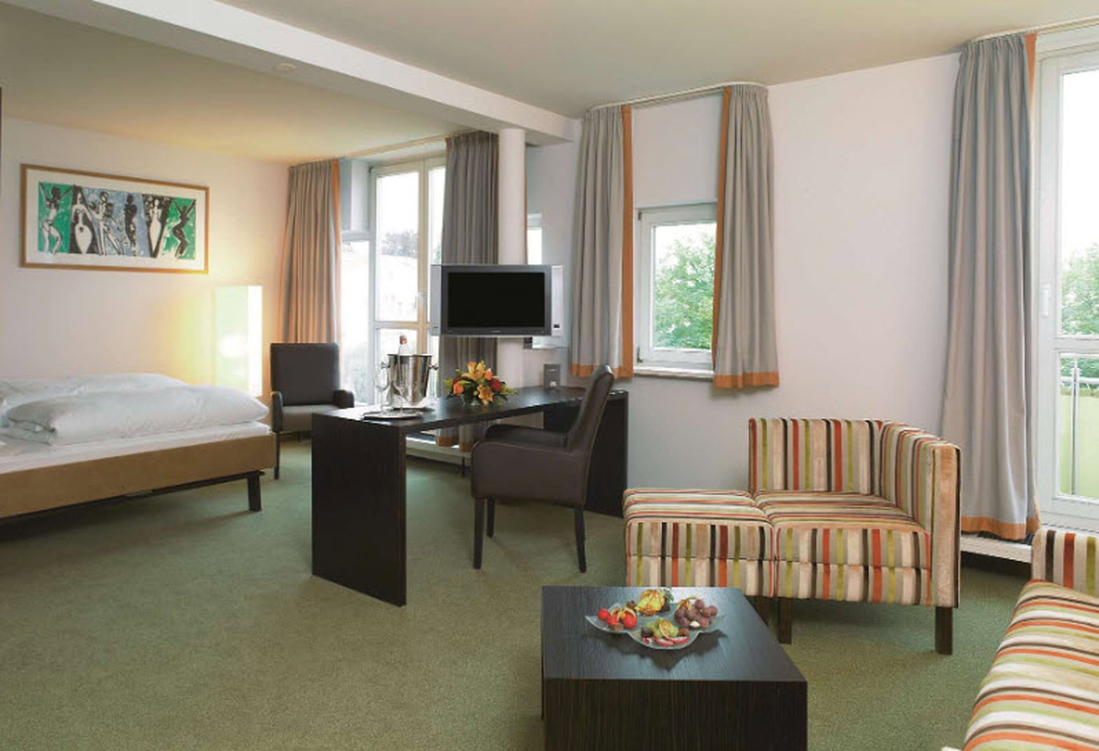 A hotel room with a bed, a desk with a TV, a striped sofa, and a dining table with fruit.