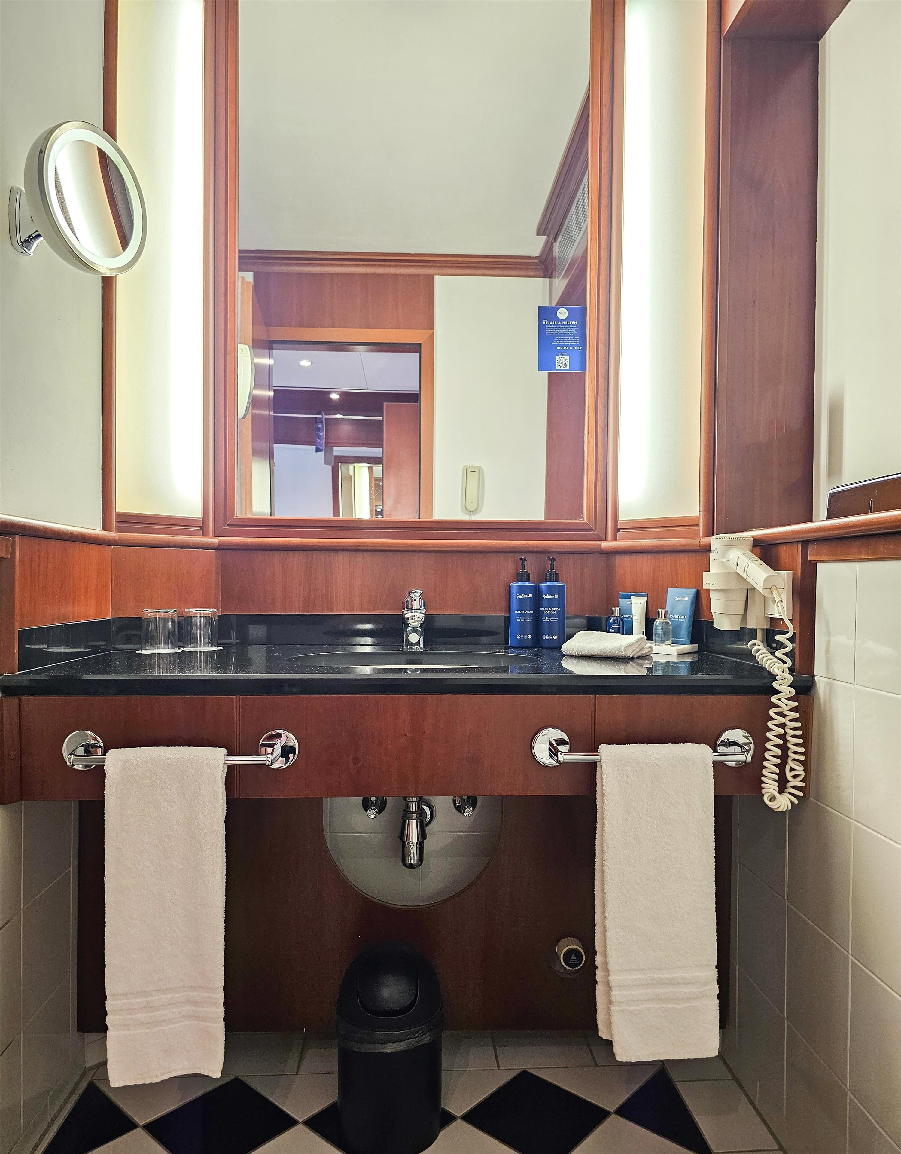A bathroom with a wooden vanity, a mirror, a sink with a black countertop, two towels, a trash can, and a telephone.