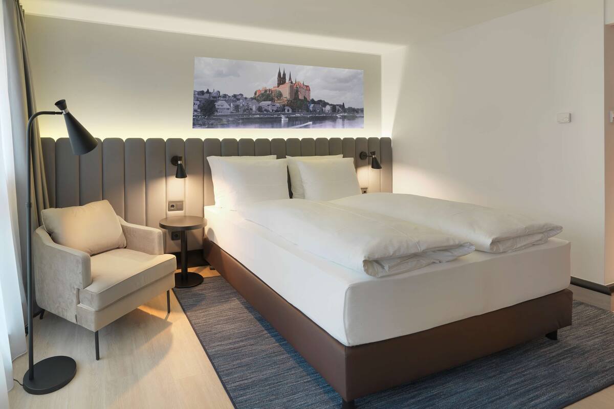 Una habitación de hotel moderna con una cama grande, una silla, una mesita y un cuadro colgado en la pared que muestra un paisaje urbano.