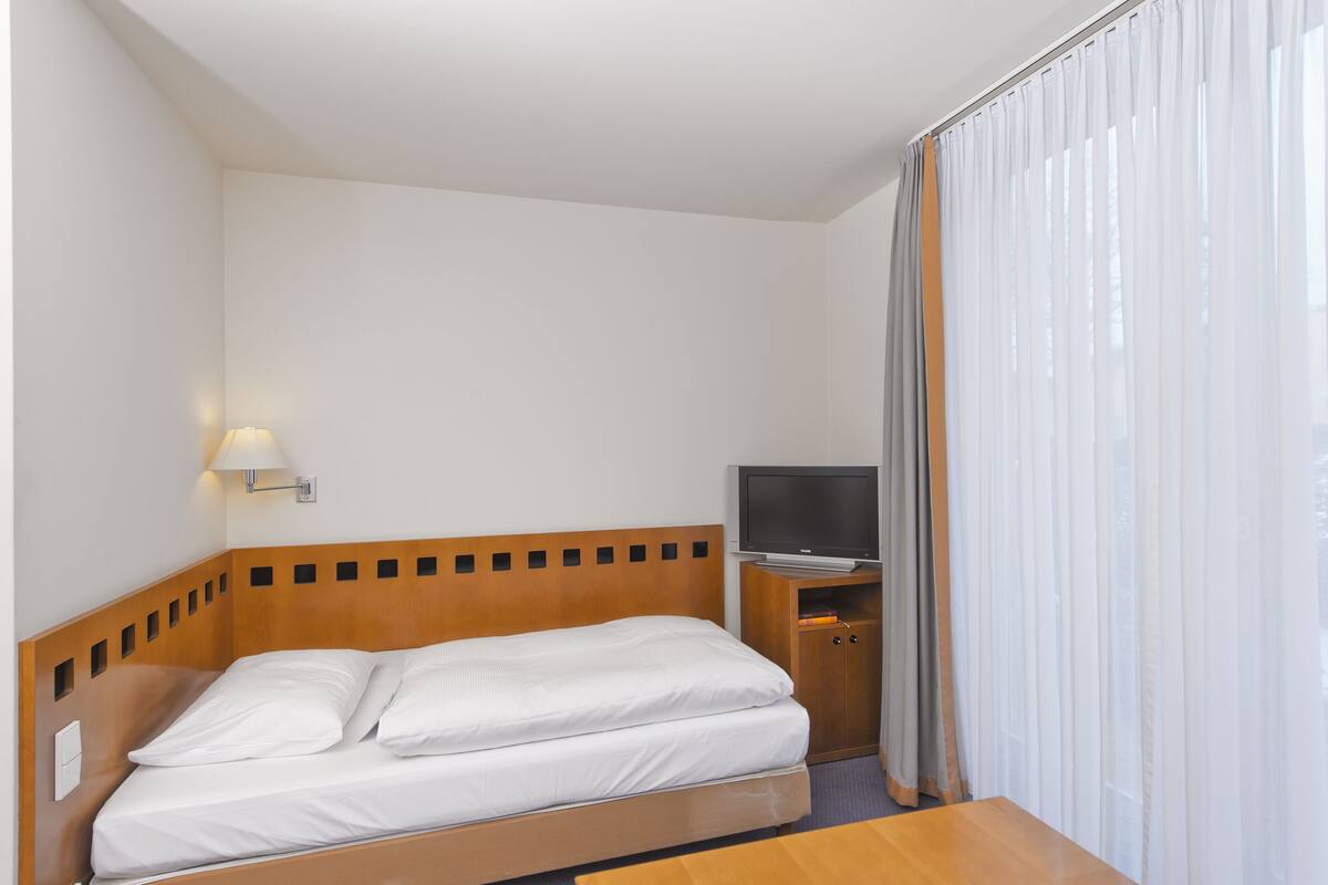 Una habitación de hotel con cabecera de madera, cama con ropa de cama blanca, televisor sobre un mueble de madera y ventana con cortinas translúcidas.