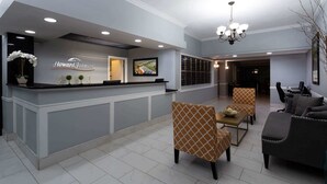 A reception area with a counter displaying the name 'Howard Johnson', a chandelier, seating with patterned upholstery, and a television screen. - Howard Johnson by Wyndham Waukegan Great Lakes (Waukegan)