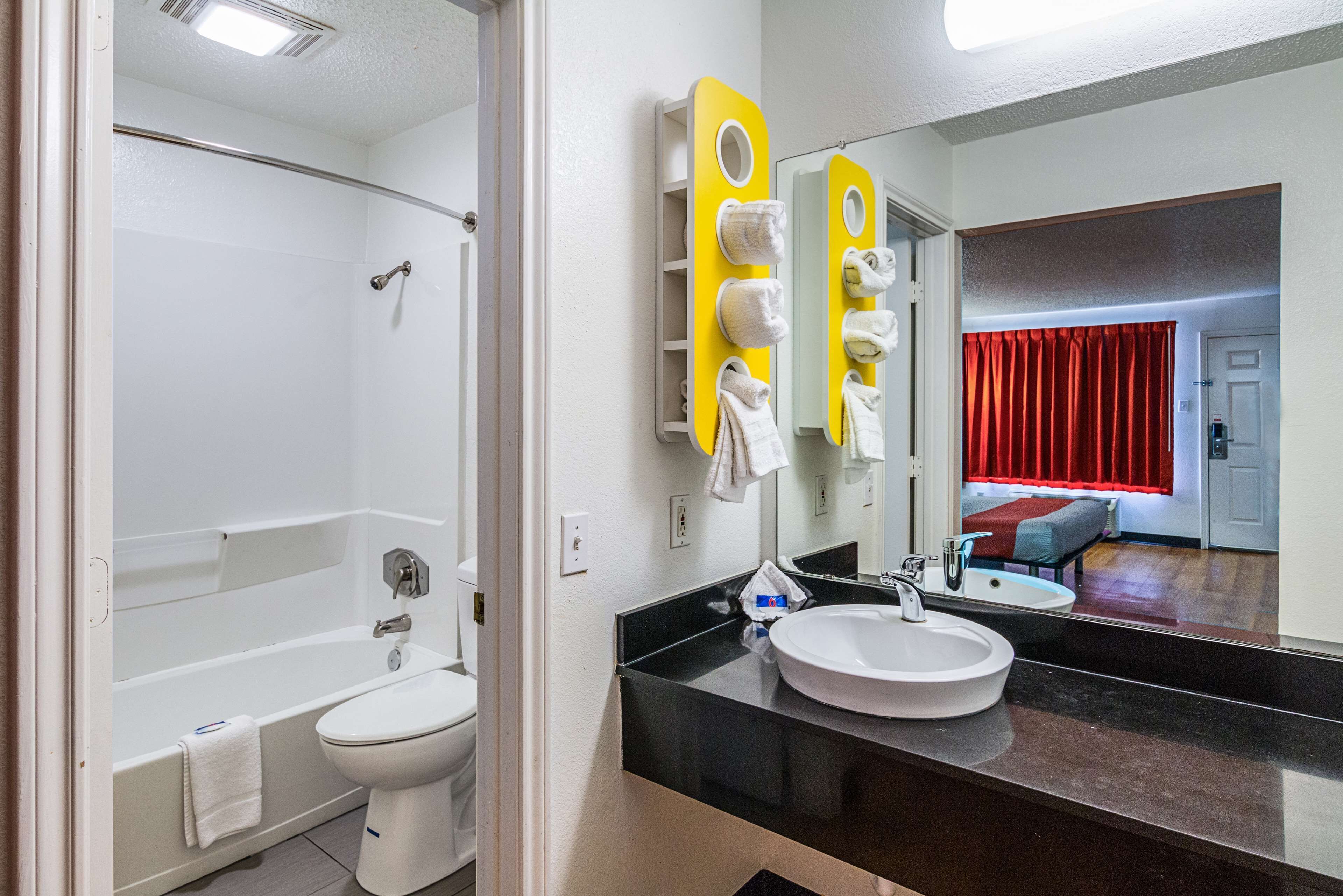 Un baño moderno con bañera, inodoro y lavamanos. Se observa un espejo con un toallero amarillo y una cortina roja al fondo.