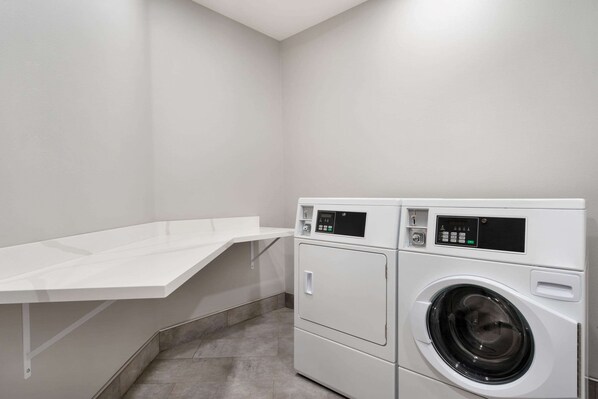 A laundry area with a white washing machine and dryer, a white shelf, and a grey tiled floor.