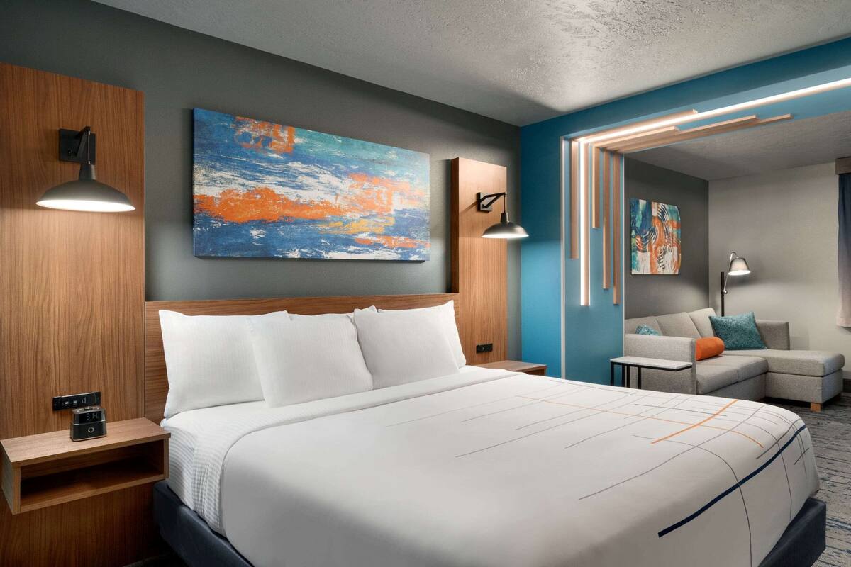 A modern hotel room with a large bed, a wooden headboard, a painting on the wall, and a seating area with a sofa and armchairs.