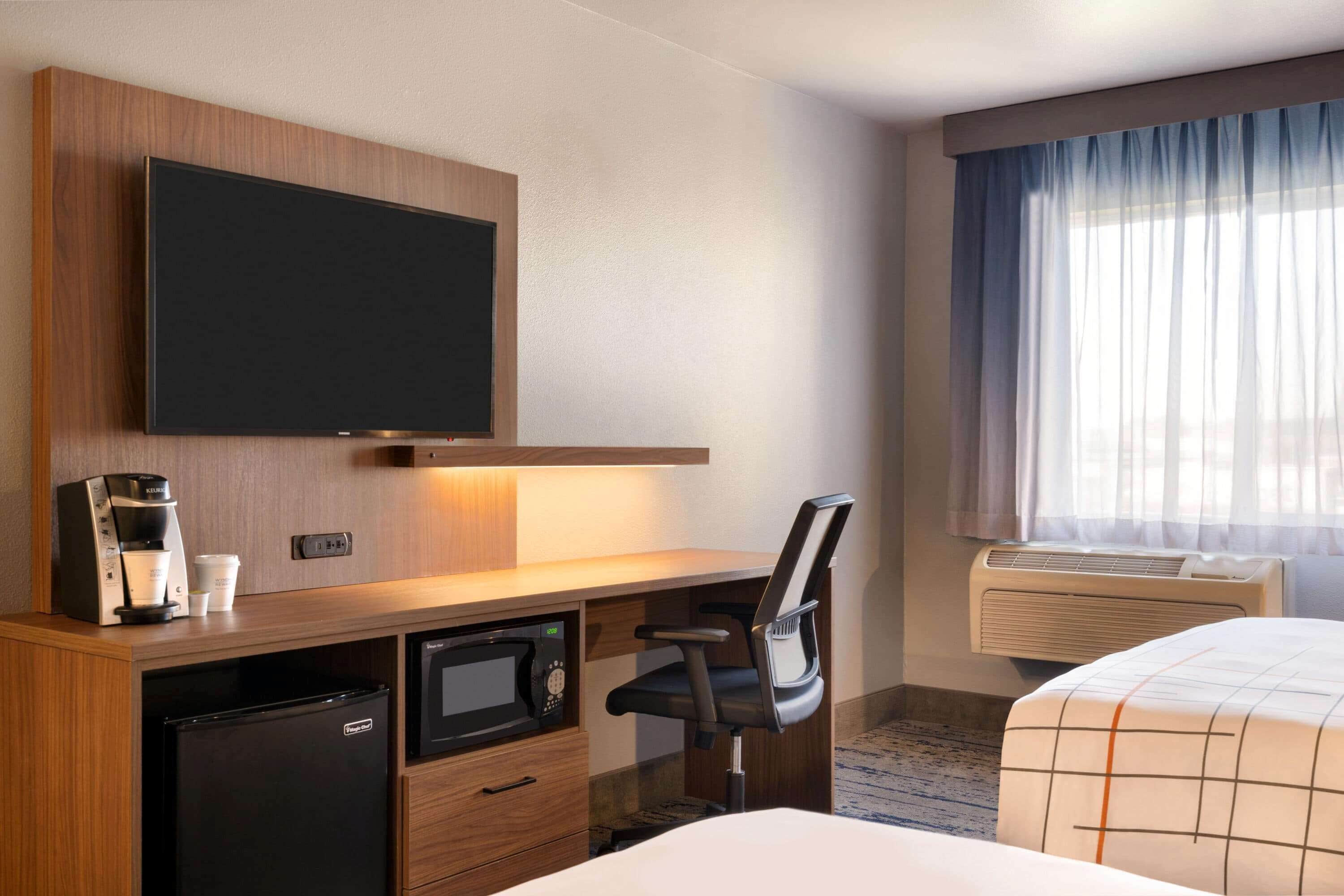 A hotel room with a flat-screen TV mounted on the wall, a desk with a coffee maker, a microwave, a chair, and a bed with a checkered bedspread.