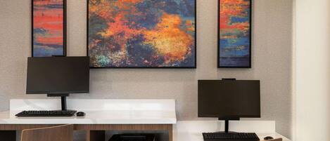 A modern office space with two desks, each with a computer monitor, keyboard, and mouse. There are three large abstract paintings on the wall above the desks.