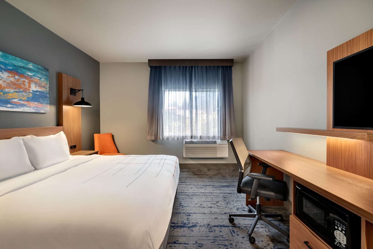 A hotel room with a large bed, a desk, an air conditioning unit, a television, and a window with curtains.