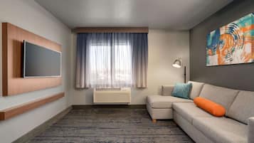 Una habitación de hotel con una televisión de pantalla plana montada en la pared, un sofá modular con un cojín naranja, una lámpara de pie y un cuadro abstracto de gran tamaño en la pared.