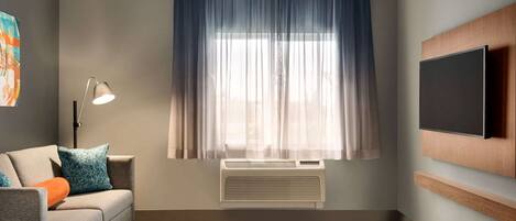 A modern hotel room with a sofa, a flat-screen TV mounted on the wall, a window with sheer curtains, and a ceiling-mounted air conditioning unit.