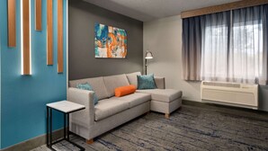 A modern living room with a sectional sofa, a small side table, a painting on the wall, and a large window with curtains. - La Quinta Inn & Suites by Wyndham Springfield (Springfield)
