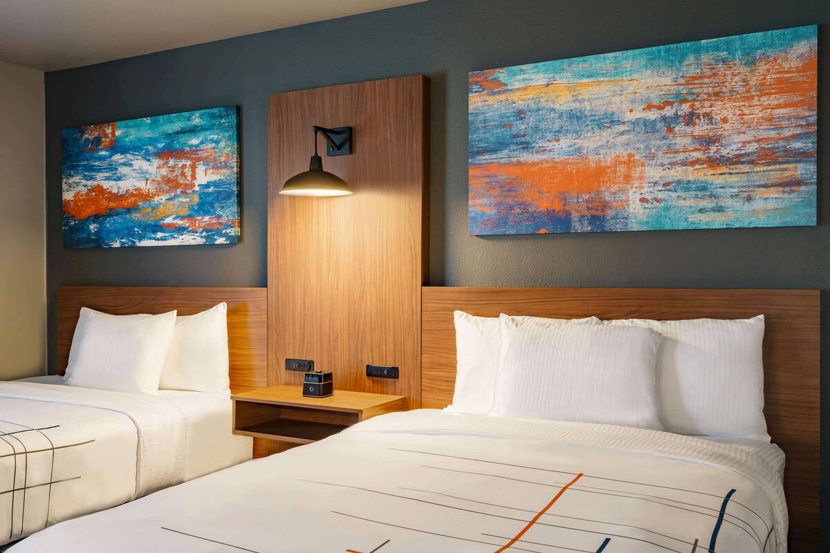A hotel room with two beds, a wooden headboard, a nightstand, and two abstract paintings on the wall.
