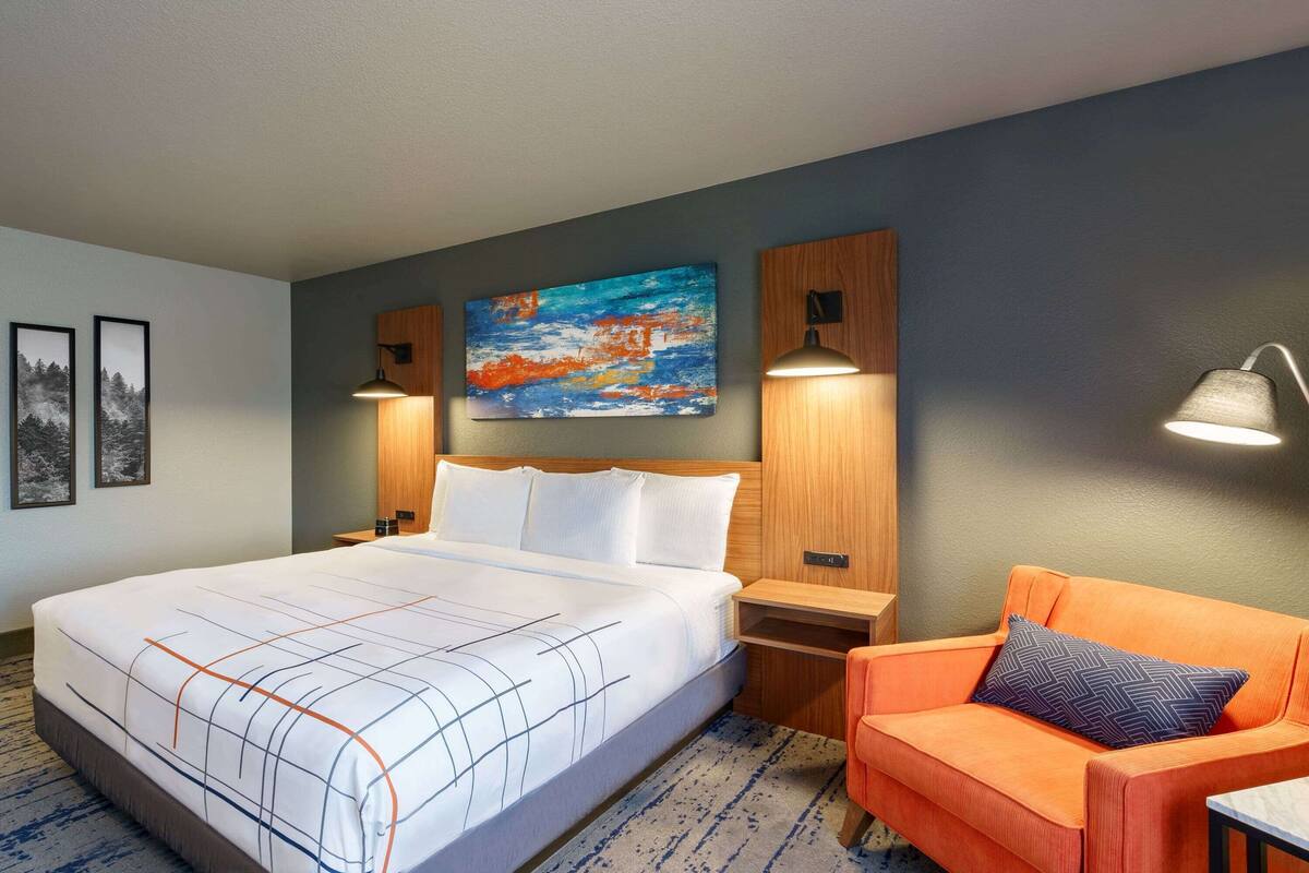 A hotel room with a large bed, a nightstand, an orange armchair, a painting on the wall, and two wall-mounted lamps.
