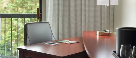A hotel room with a wooden desk, a chair, a lamp, and a view of trees through the window.