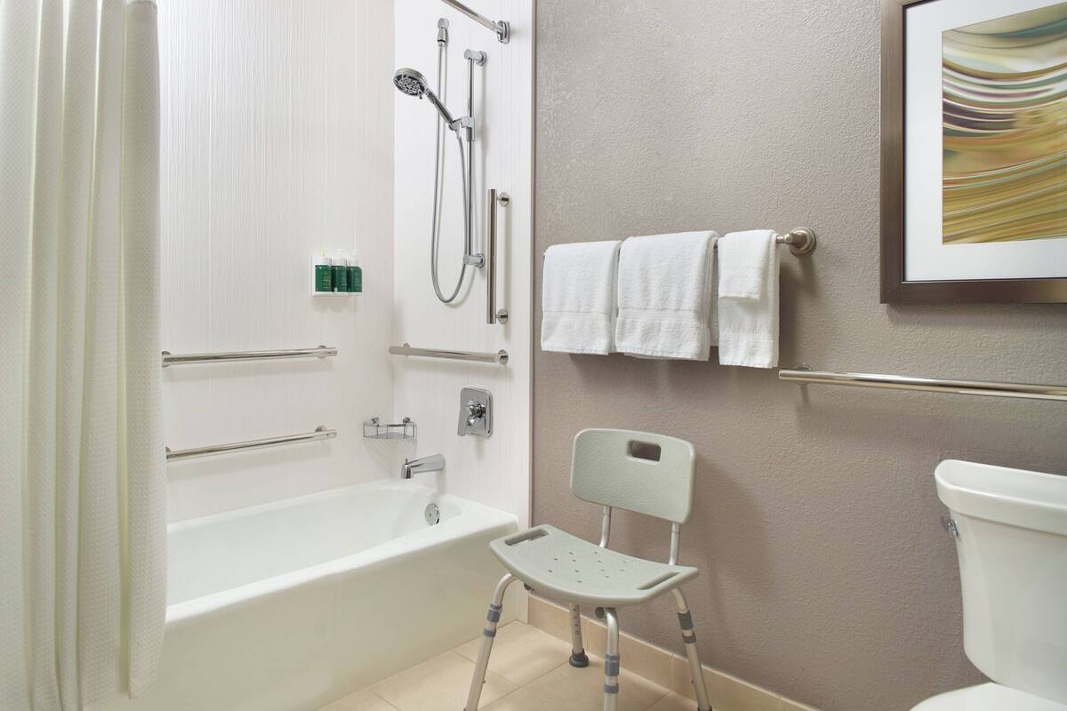A bathroom with a bathtub, shower, grab bars, a toilet, and a chair.