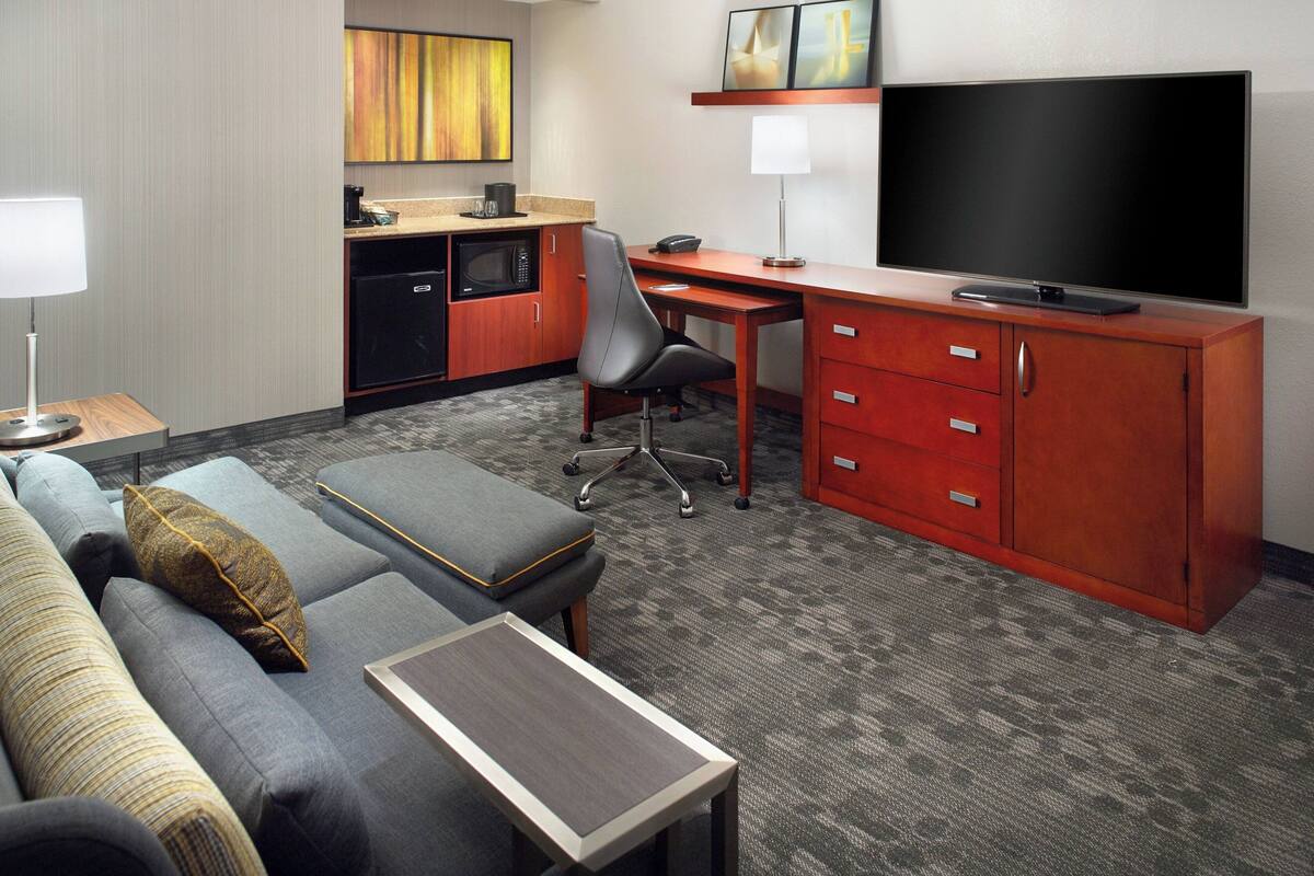 A hotel room with a sofa, ottoman, desk, TV, kitchenette, and a small table.