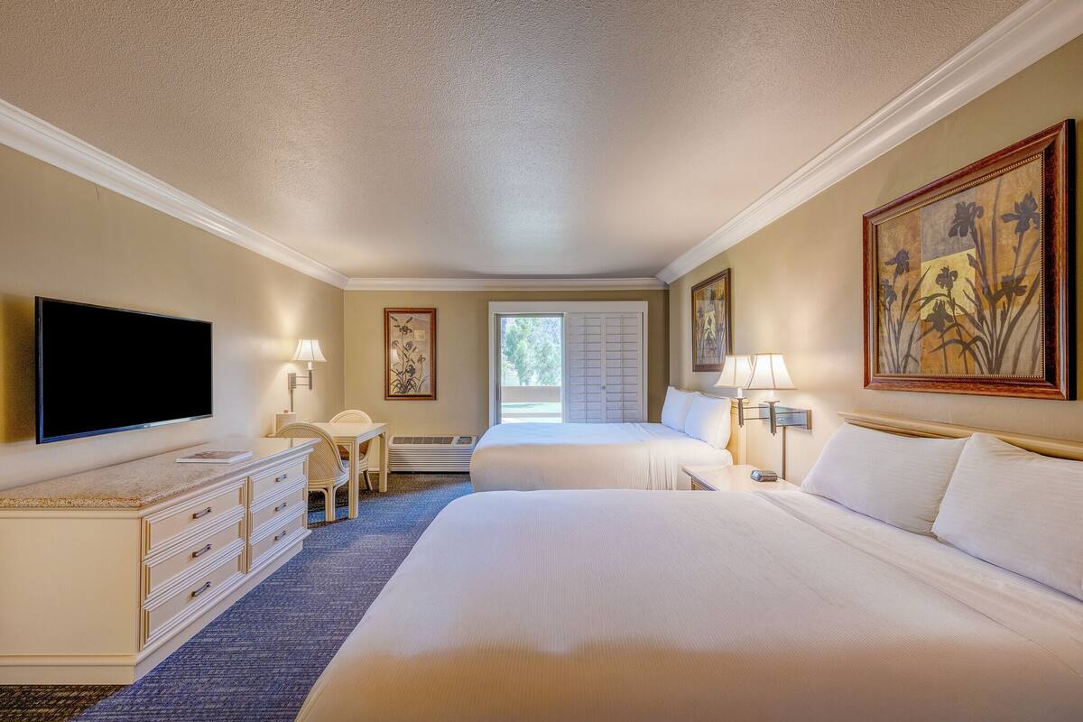 A hotel room with two beds, a TV, a desk, and a window with blinds.