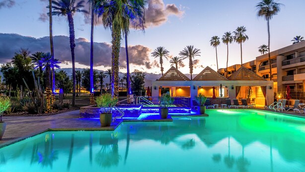 A resort pool area with palm trees, a pool, and a building with a thatched roof. - Indian Wells Resort Hotel (Indian Wells)
