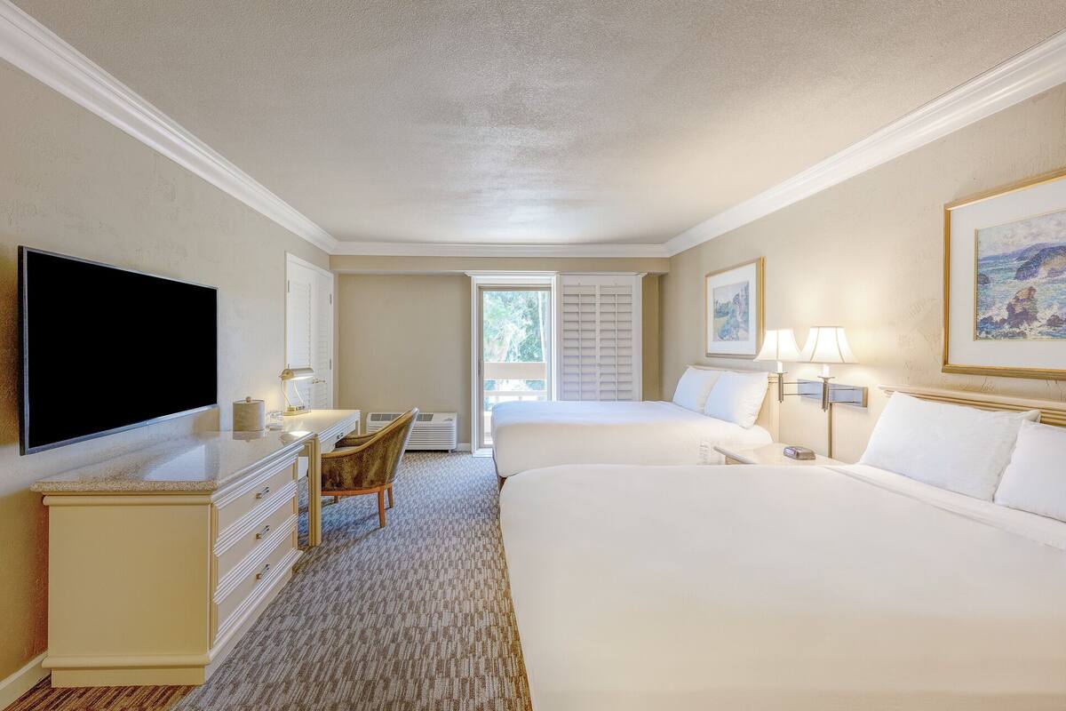 A hotel room with two beds, a desk with a flat-screen TV, a chair, and a window with blinds.