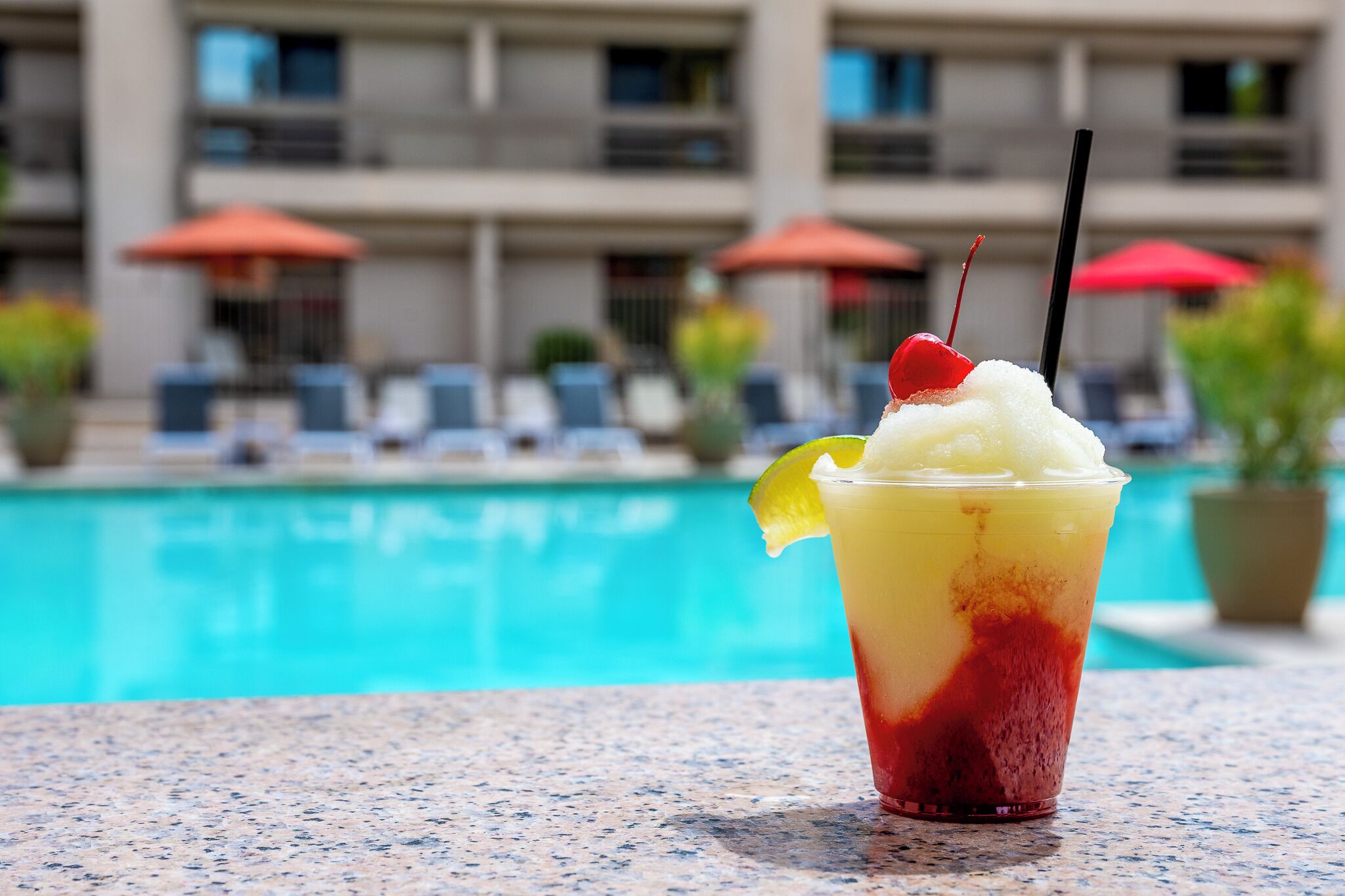 En avslappnad miljö vid poolkanten med en drink i ett glas, en pool och en byggnad med balkonger och parasoller i bakgrunden.