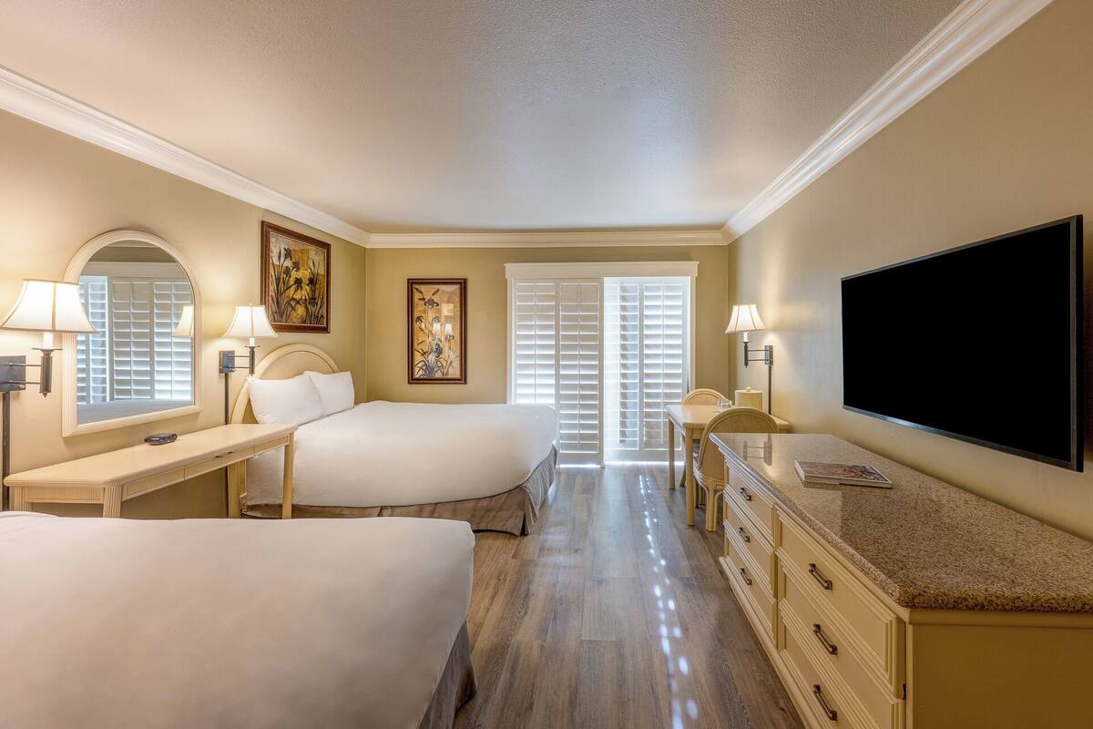 A hotel room with two beds, a television, a desk with a lamp, a chair, and a large window with blinds.
