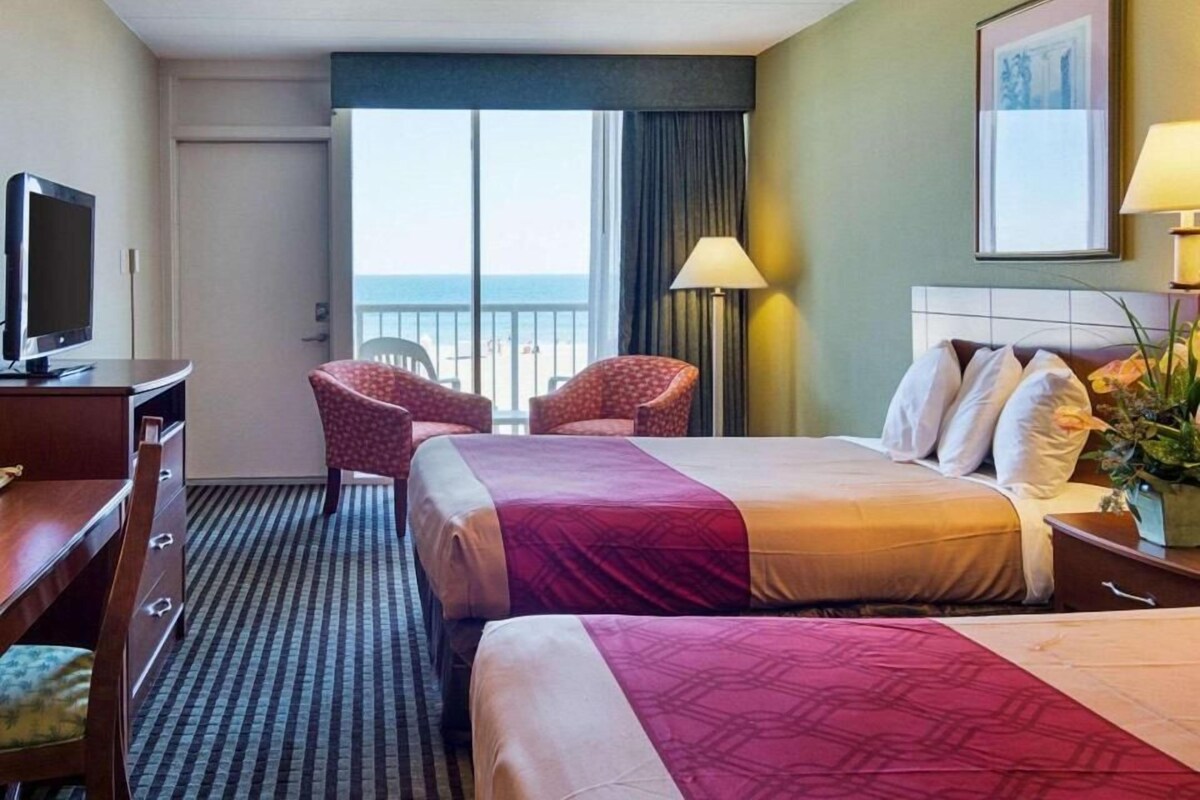 A hotel room with two beds, a desk, a chair, a TV, and a balcony with a view.
