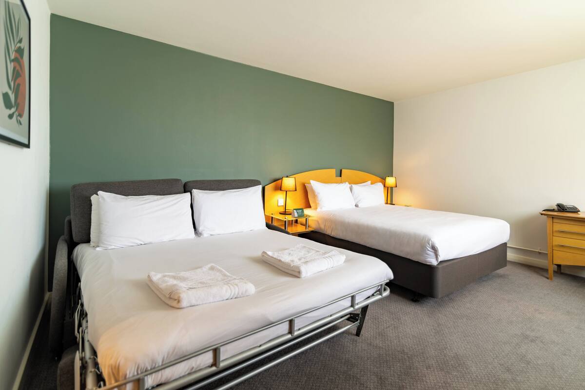 A hotel room with two beds, each with white linens and pillows, a bedside table with a lamp, and a small wooden nightstand.