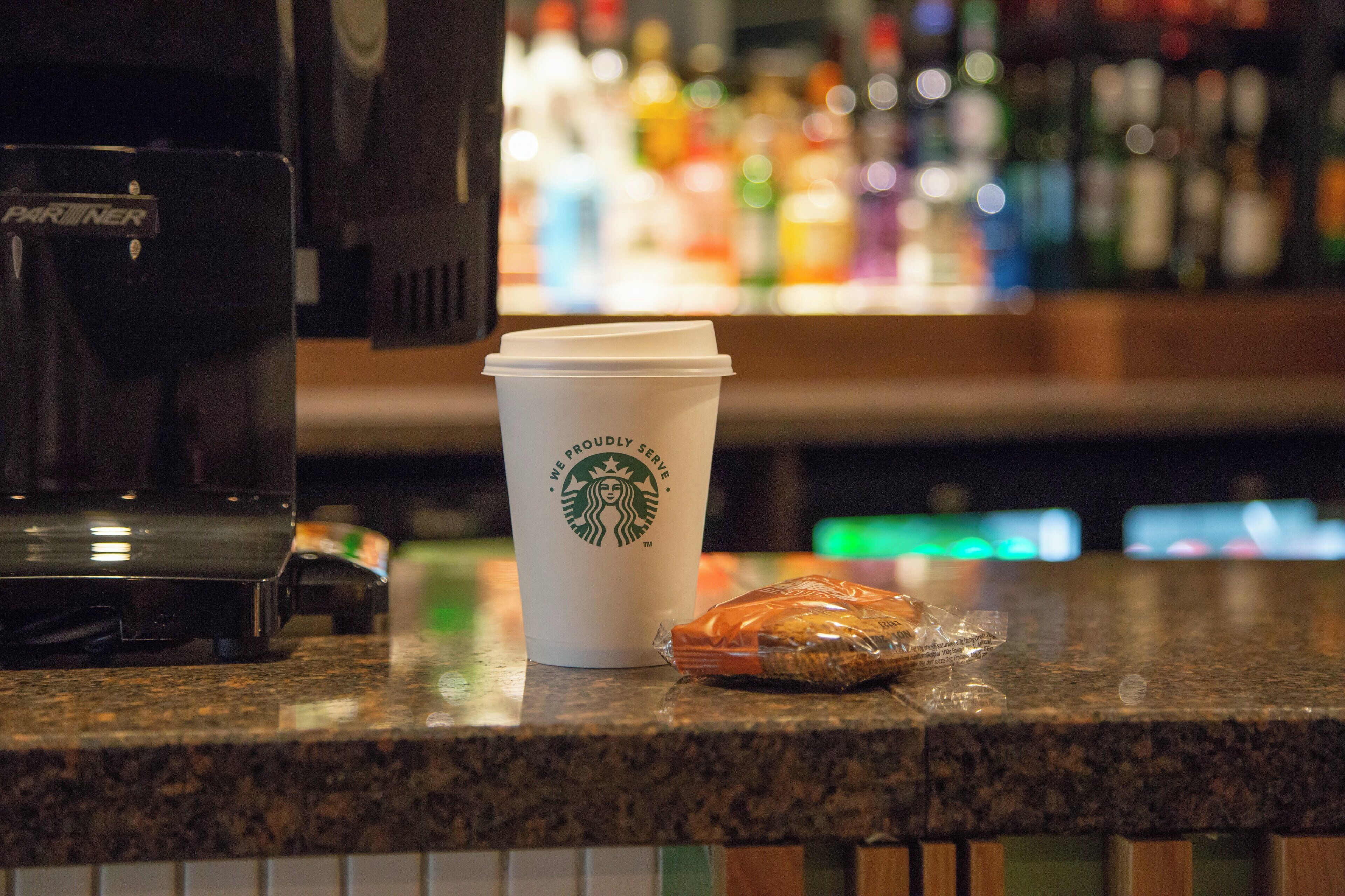 A Starbucks cup and a pastry on a counter with a coffee machine and a blurred background of bottles.
