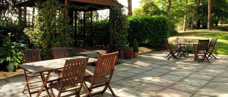 An outdoor patio with a wooden table and chairs, surrounded by potted plants and a pergola.