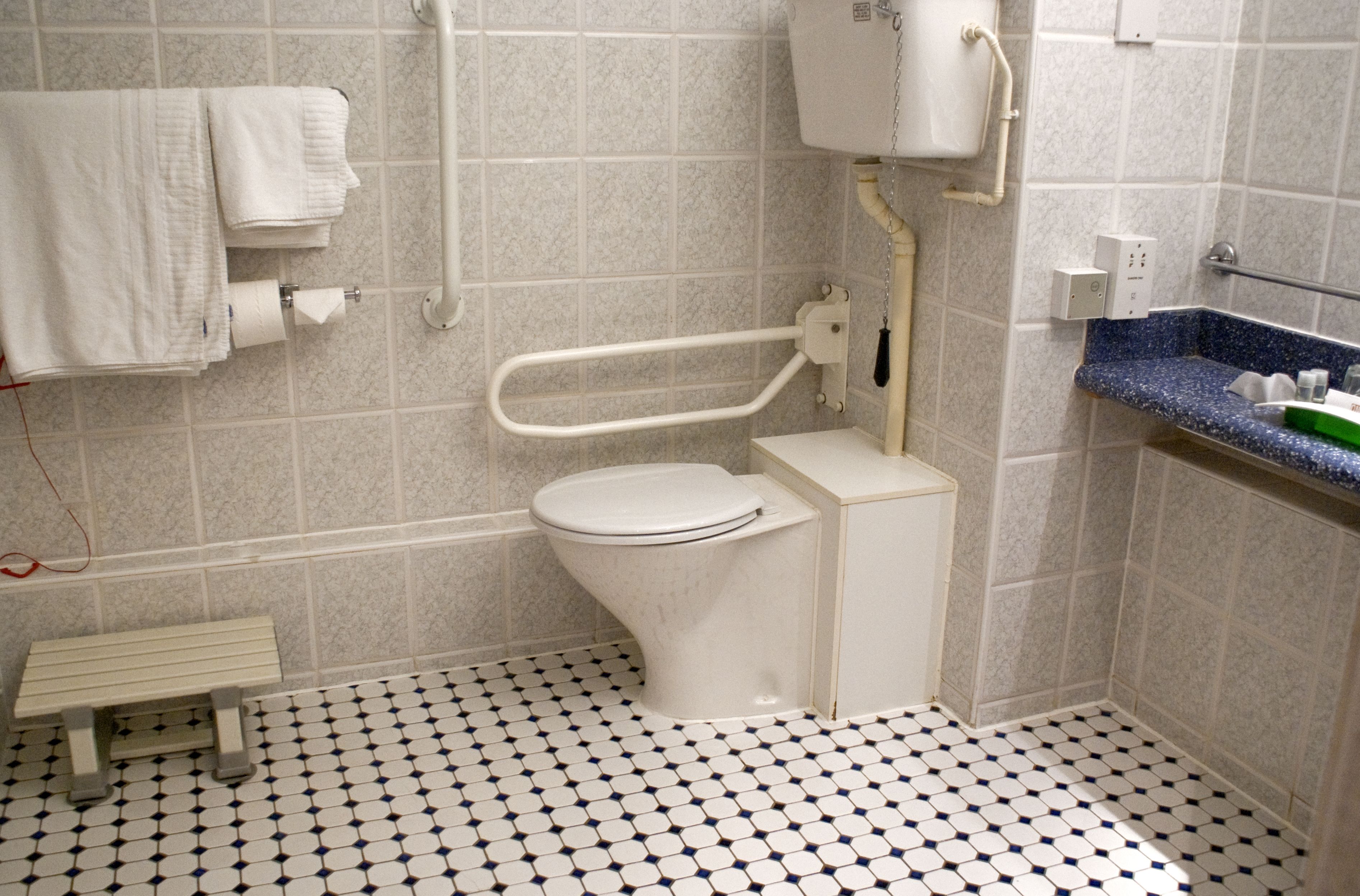 A bathroom with a white toilet, a bench, a sink with a blue countertop, and a tiled floor.