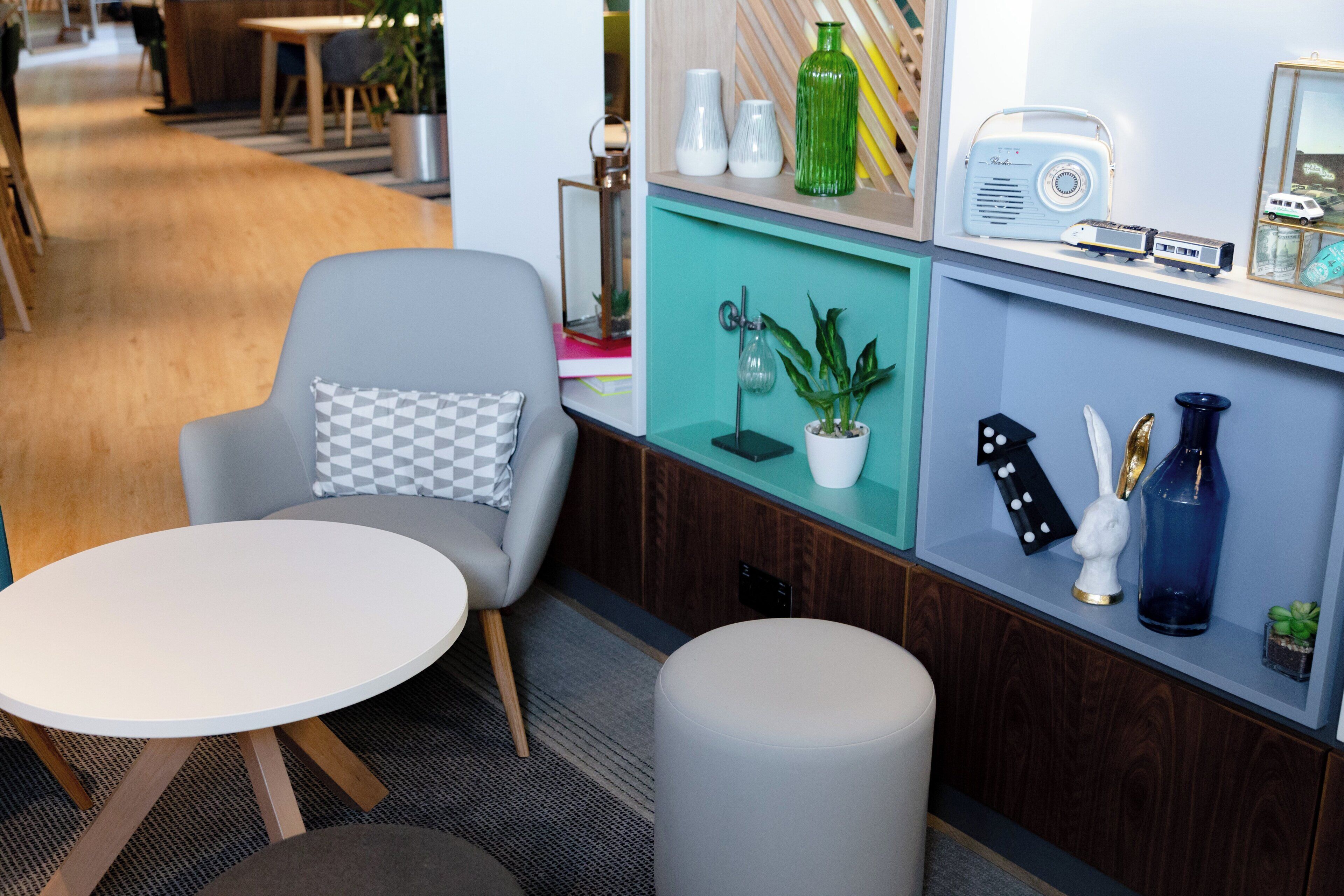 Ein modernes Interieur mit einem grauen Sessel, einem runden weißen Tisch und einem kleinen Hocker. Regale mit Dekorationsobjekten und ein Radio sind ebenfalls zu sehen.