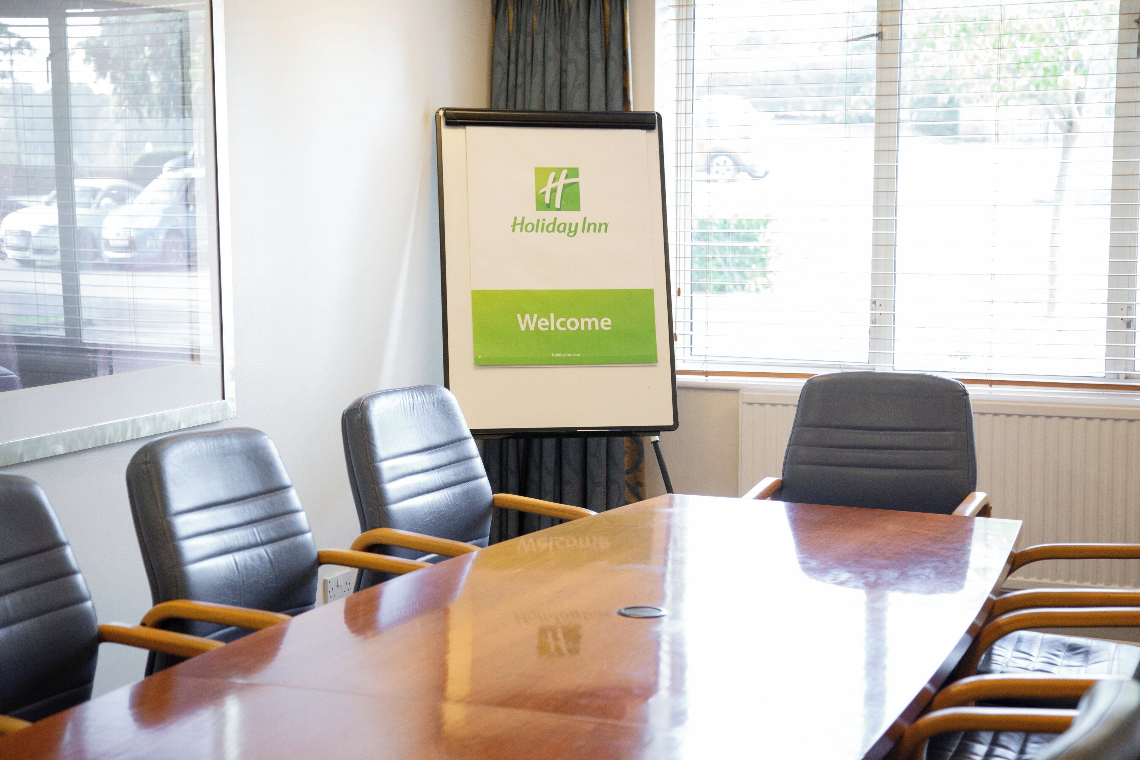 A conference room with a wooden table, four leather chairs, and a whiteboard with 'Holiday Inn Welcome' written on it.