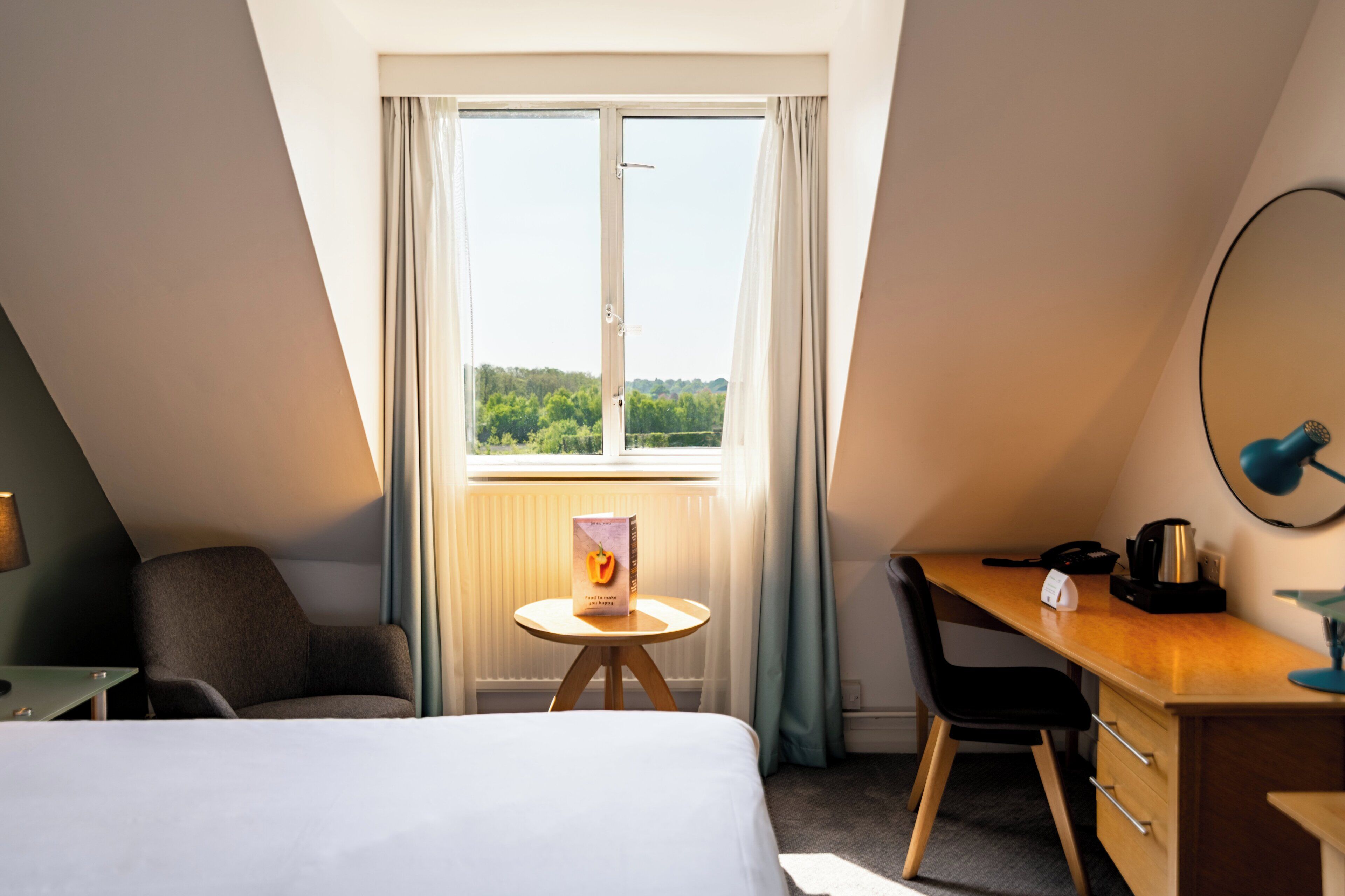 Ein Hotelzimmer mit Bett, Schreibtisch, Stuhl, einem kleinen Tisch und einem Fenster mit Vorhängen.