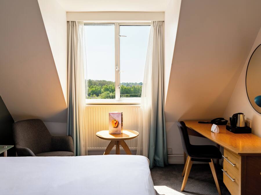 Ein Hotelzimmer mit Bett, Schreibtisch, Stuhl, einem kleinen Tisch und einem Fenster mit Vorhängen.