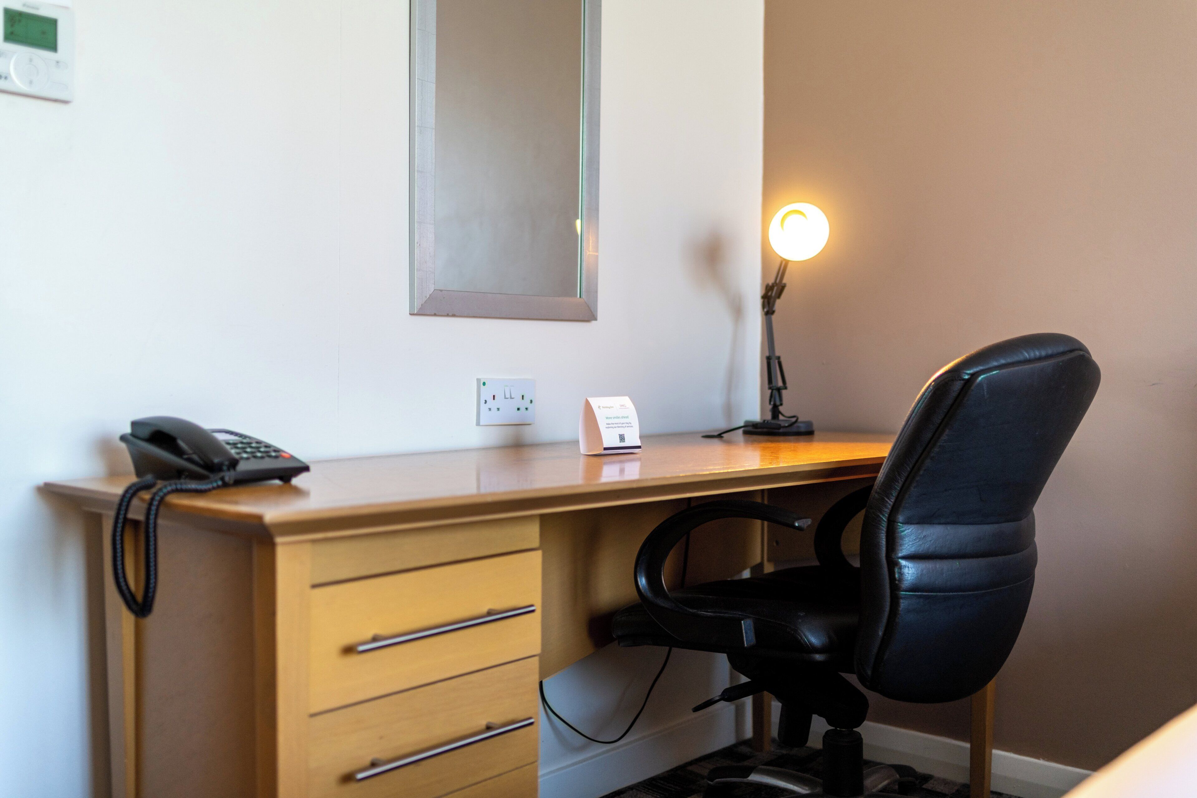 Ein Hotelzimmer mit einem Holztisch, einem schwarzen Bürostuhl, einer Schreibtischlampe, einem Telefon und einem an der Wand befestigten Spiegel.