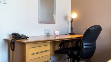 Habitación de hotel con un escritorio de madera, una silla de oficina negra, una lámpara de escritorio, un teléfono y un espejo montado en la pared.