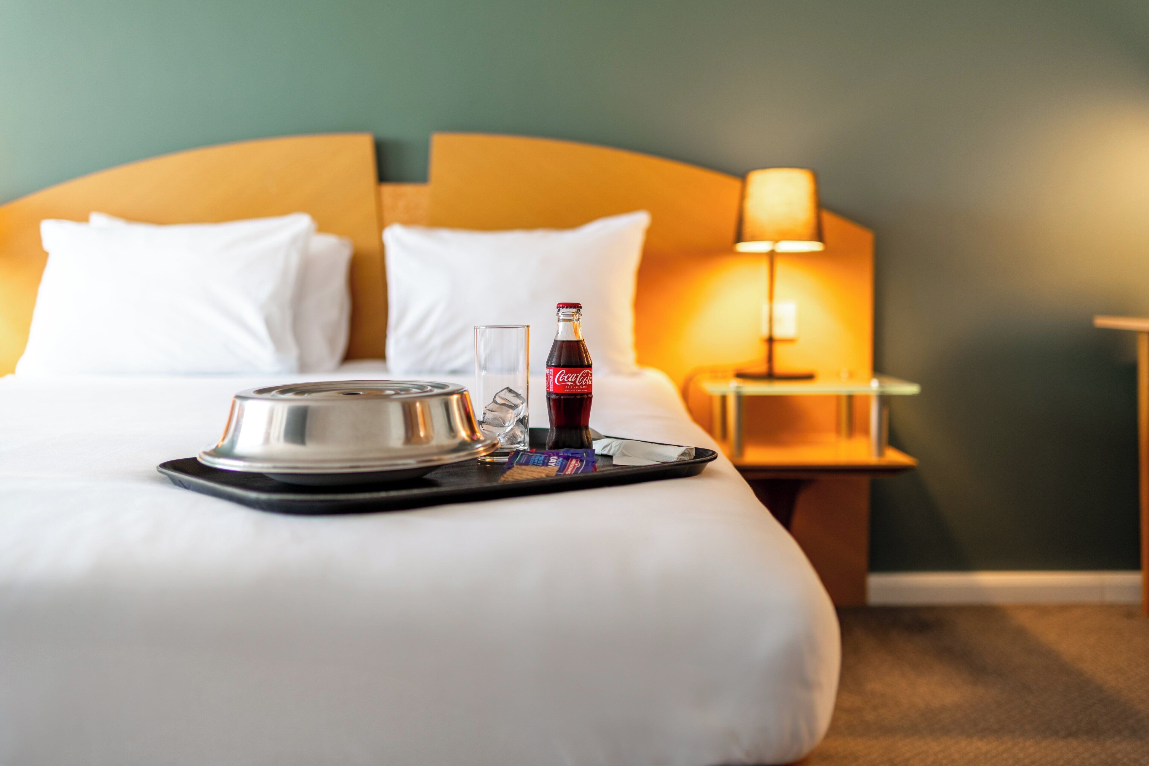Una cama bien hecha con almohadas blancas y una bandeja con una botella de Coca-Cola, un vaso y un plato pequeño.