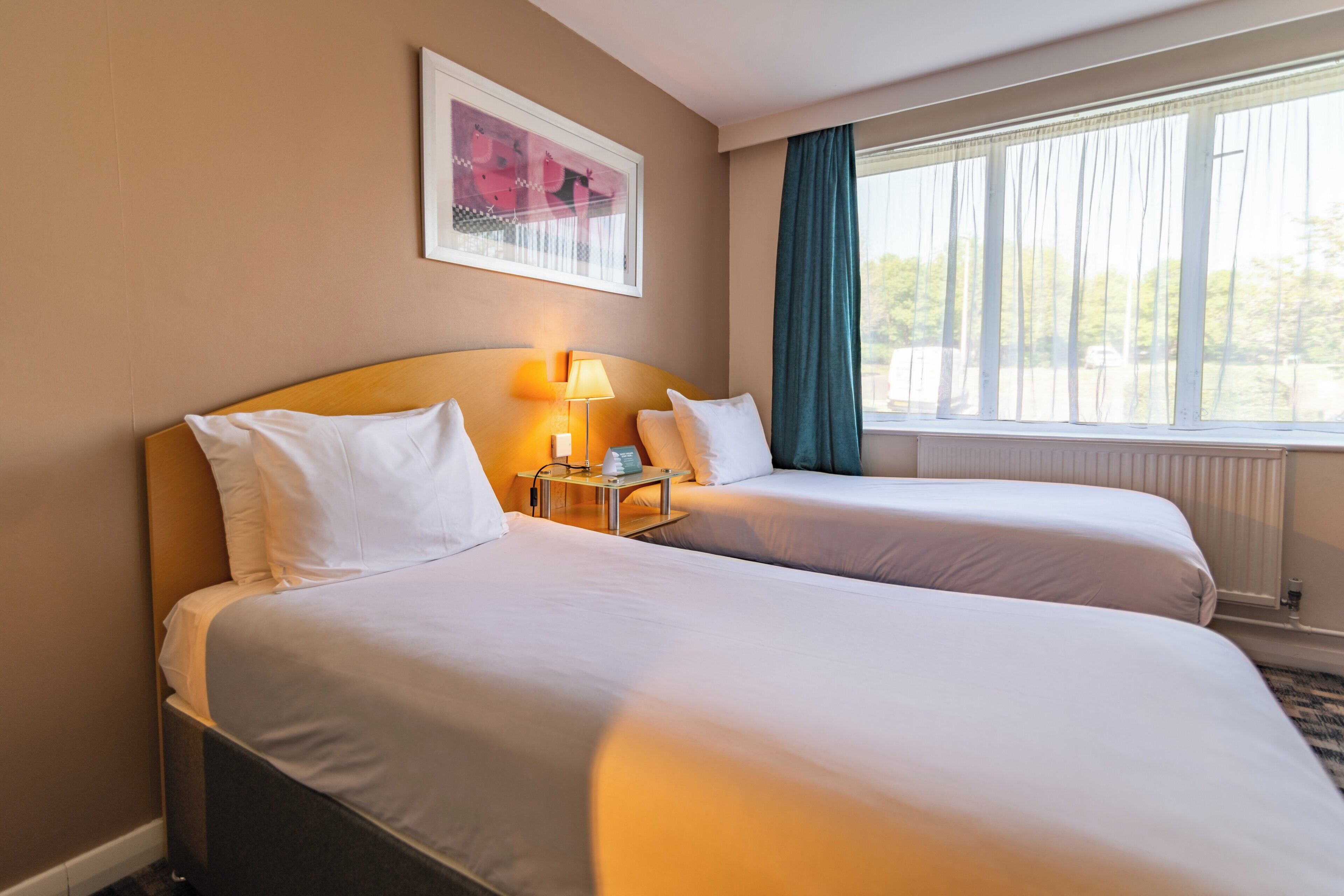 A hotel room with two beds, a nightstand with a lamp, a framed picture on the wall, and a window with curtains.