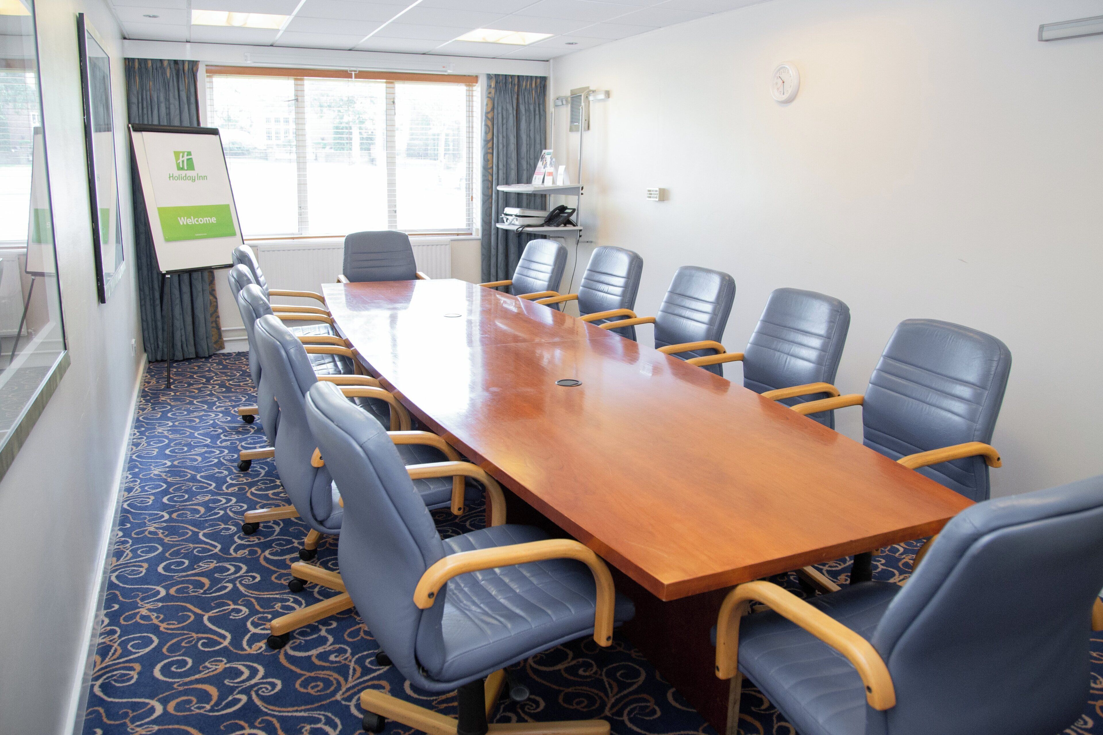 A conference room with a long table and multiple chairs, a clock on the wall, and a window with blinds.
