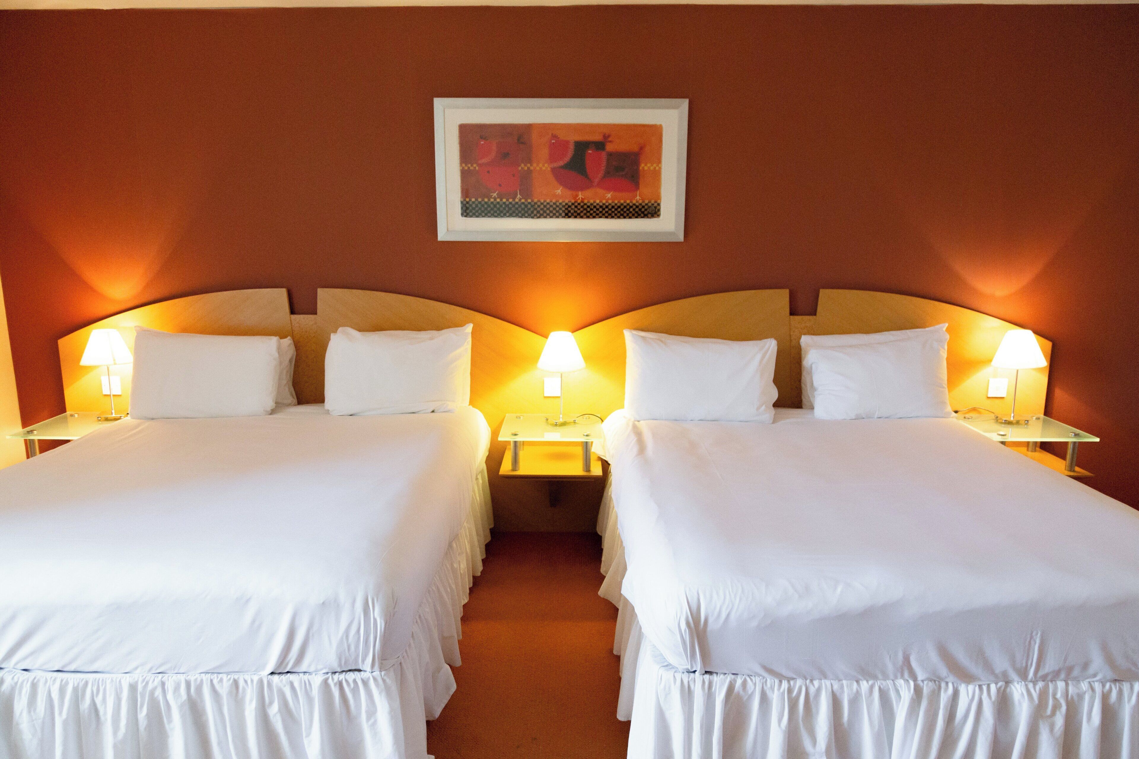 A hotel room with two beds, each with a white bedspread and pillows, flanked by nightstands with lamps. A framed artwork hangs on the wall above the beds.