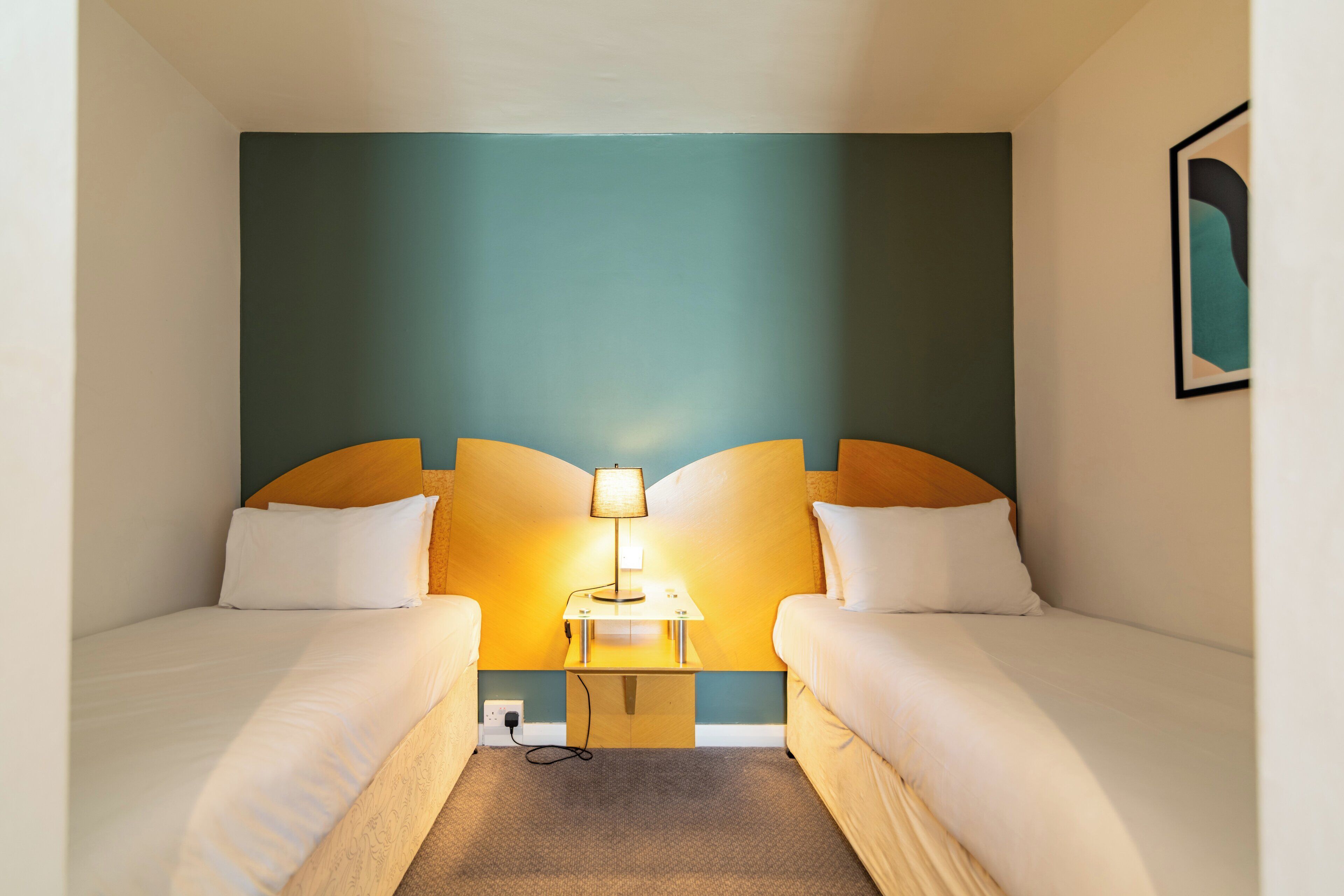 A hotel room with two single beds, a bedside table with a lamp, and a wall-mounted artwork.
