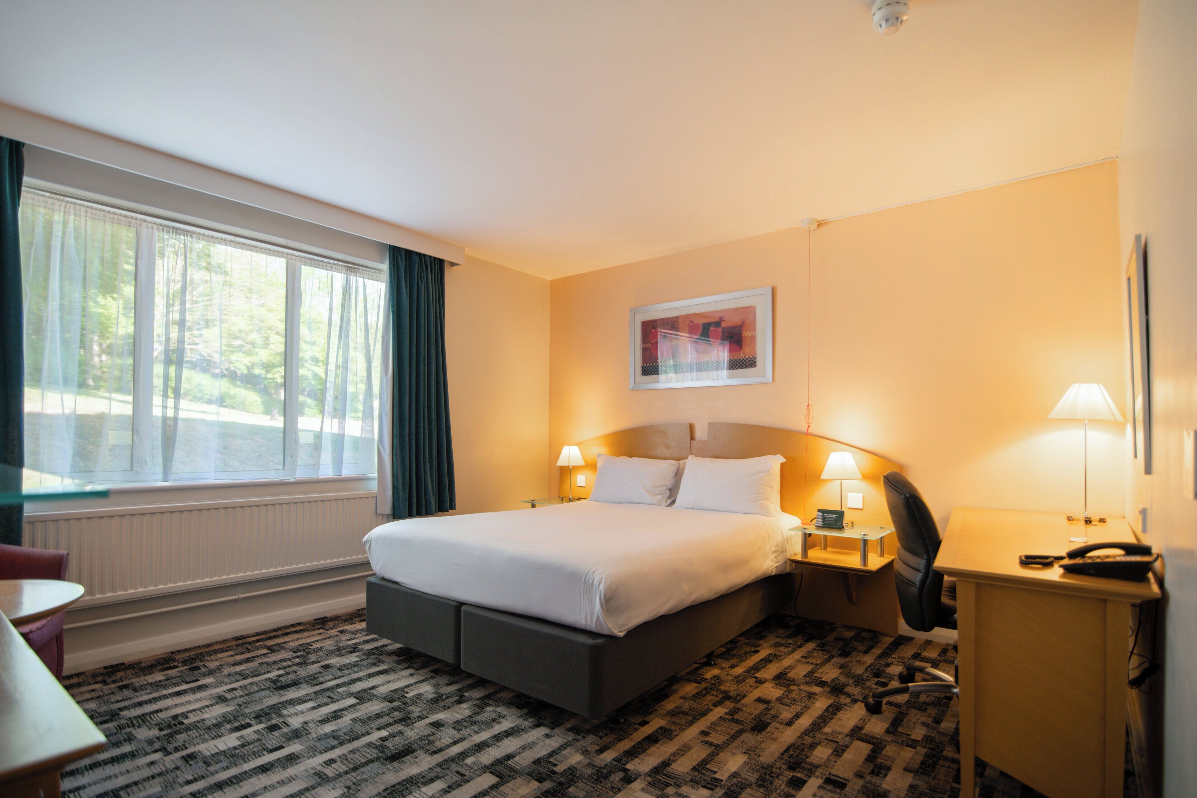 Une chambre d’hôtel comprenant un lit, des tables de chevet, un bureau, une chaise, une fenêtre avec des rideaux et un radiateur.