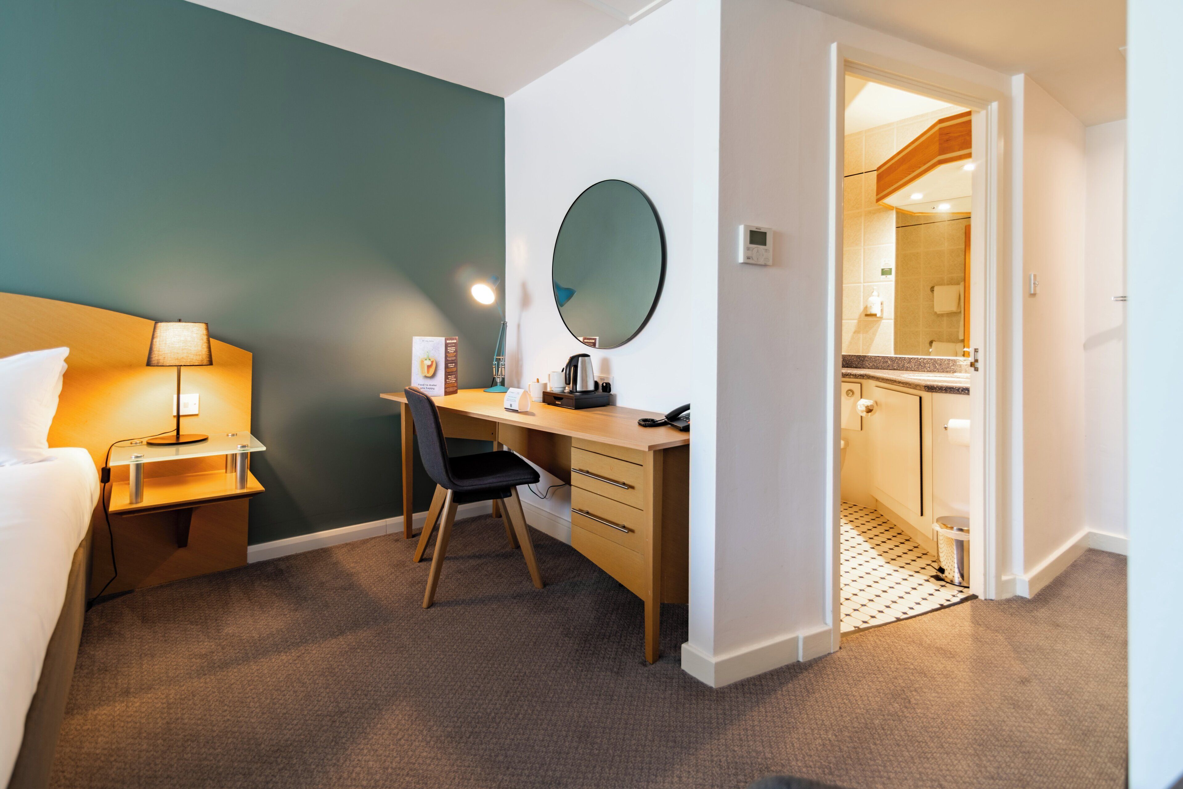 Ein Hotelzimmer mit Bett, Schreibtisch, Stuhl und einem großen Spiegel. Ein Badezimmer ist durch eine offene Tür sichtbar.